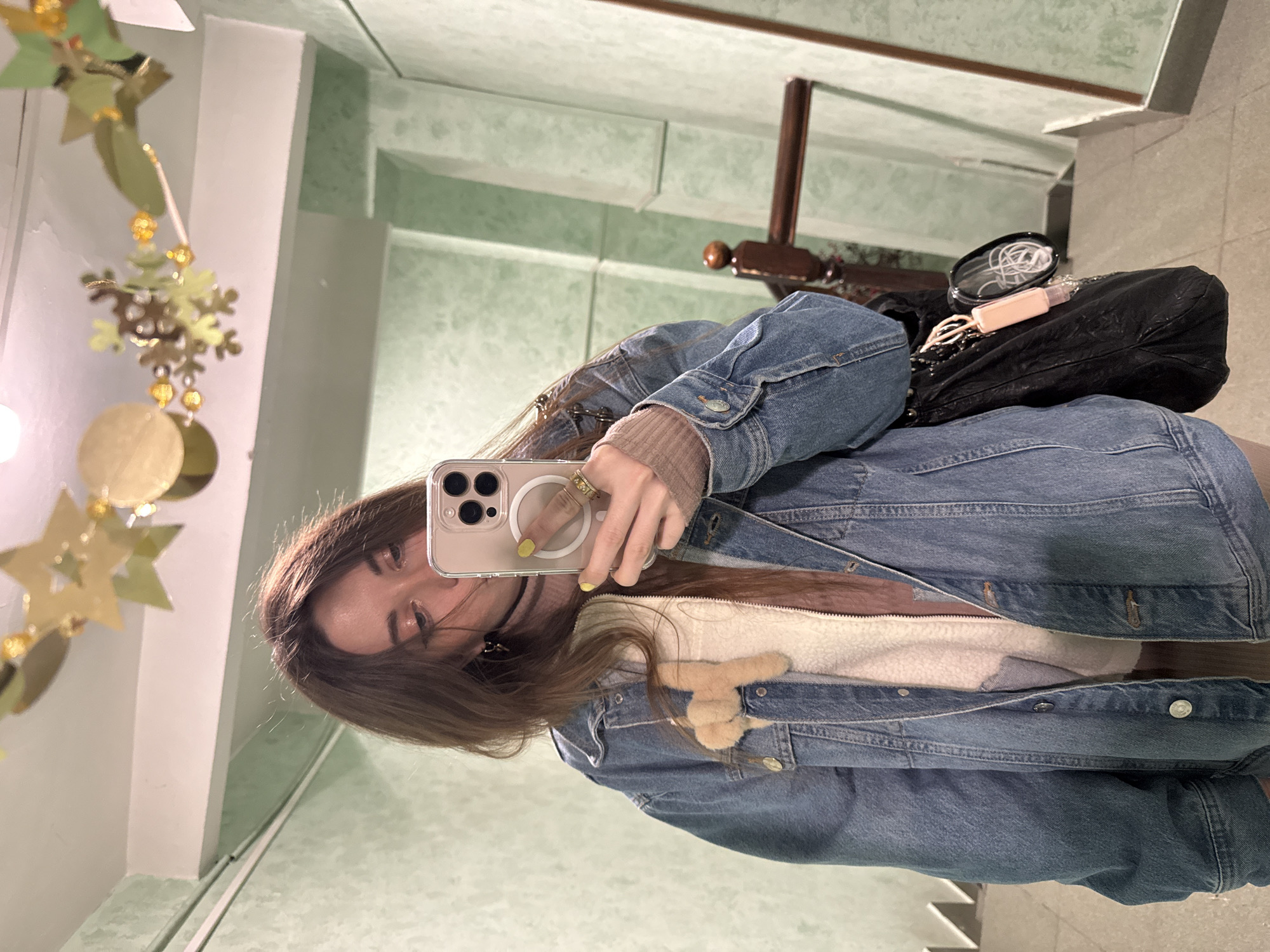 A person is taking a selfie in a mirror inside a building with green walls and white trim. The person is wearing a denim jacket over a light-colored top with a fuzzy embellishment. The person has long brown hair and is holding a phone with a ring magnet on the back. In the background, there is a dark purse and a decorative hanging ornament with stars and circles. The location is Moscow, Russia. There is a staircase in the background.