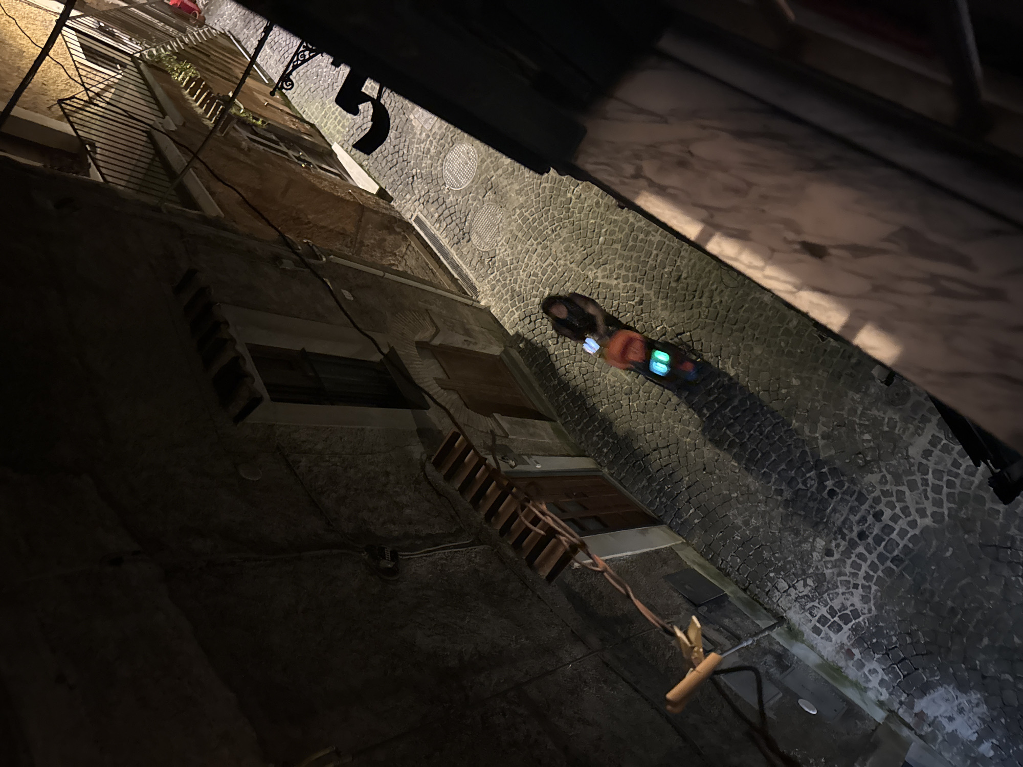 The image shows a view of a narrow street in Castelnuovo di Porto, Italy, at night. The cobblestone street is lit dimly, reflecting light from an unidentified source. Pedestrians are walking on the street. Buildings line the street, and a ladder is visible extending from one of the buildings. There are objects in the sky, but they are obscured.