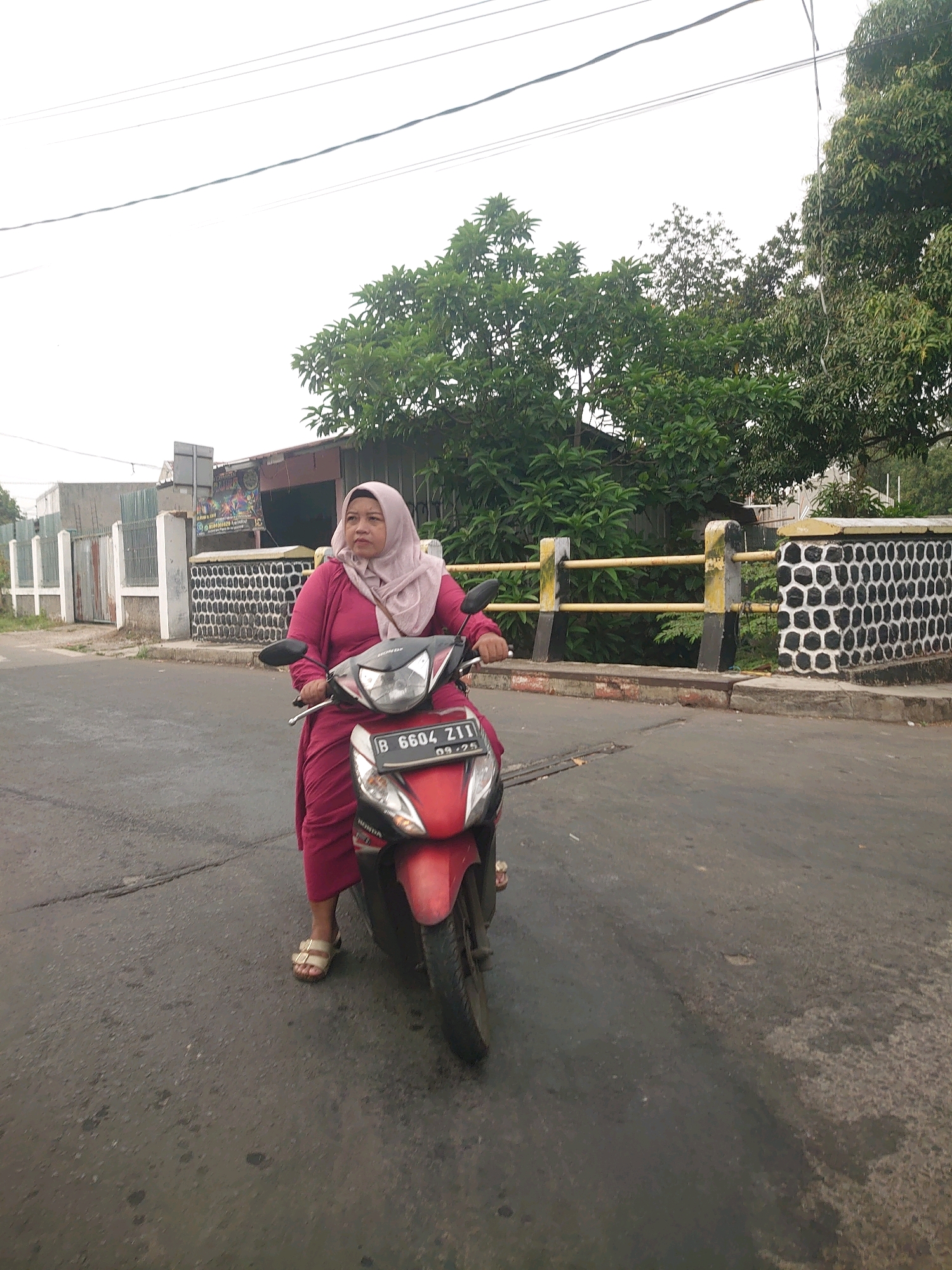 A woman wearing a pink hijab and a pink dress rides a red scooter. She is on a street in Depok, Indonesia. The street is paved and has some cracks and patches. On the left side of the street, there is a fence and a building. On the right side of the street, there is a wall with a decorative pattern and some trees. The sky is overcast. The scooter has a license plate that reads "B 6604 ZII".