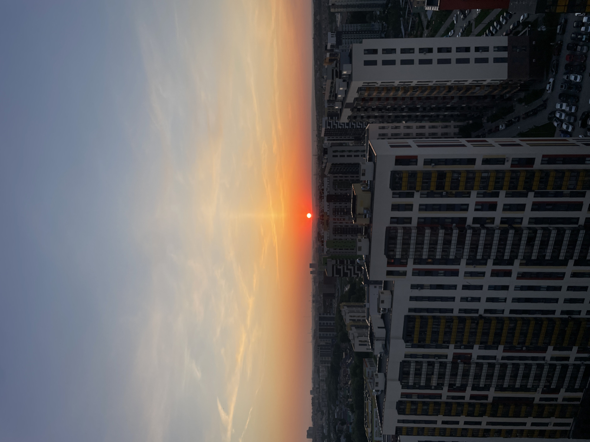 The image depicts a cityscape at sunset. The sky transitions from light blue to orange and yellow as it approaches the horizon, where the sun appears as a red disk. Below the horizon, a dense urban area is visible, featuring apartment buildings, roads, and vehicles. The structures have a modern aesthetic with repeating patterns of windows and balconies. The scene is captured from an elevated perspective, possibly a rooftop or high floor of a building. The location is in the city of Kazan, Russia.