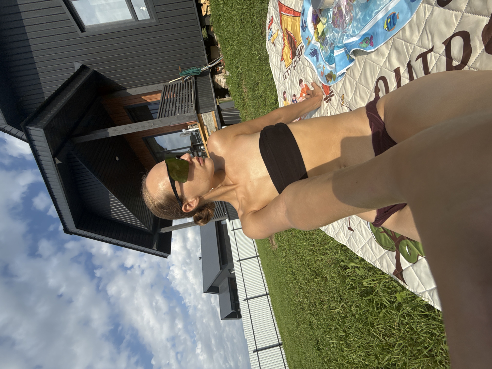 A young woman wearing a black strapless bikini top and dark red bottoms is lying on a patterned blanket on a grassy lawn. She is wearing large, mirrored sunglasses and has her hair pulled back in a bun. Her left arm is extended towards the camera, holding it in a selfie position. Behind her, a modern, dark-colored house with a prominent pitched roof stands on a slightly elevated area. A white metal fence runs horizontally across the middle ground, separating the lawn from what appears to be more houses in the distance. The sky is partly cloudy with bright sunlight, indicating it is daytime. The overall setting is a residential area, likely a backyard or a rural property, given the presence of a house and lawn. The location is identified as Podshivalovo, Russia.