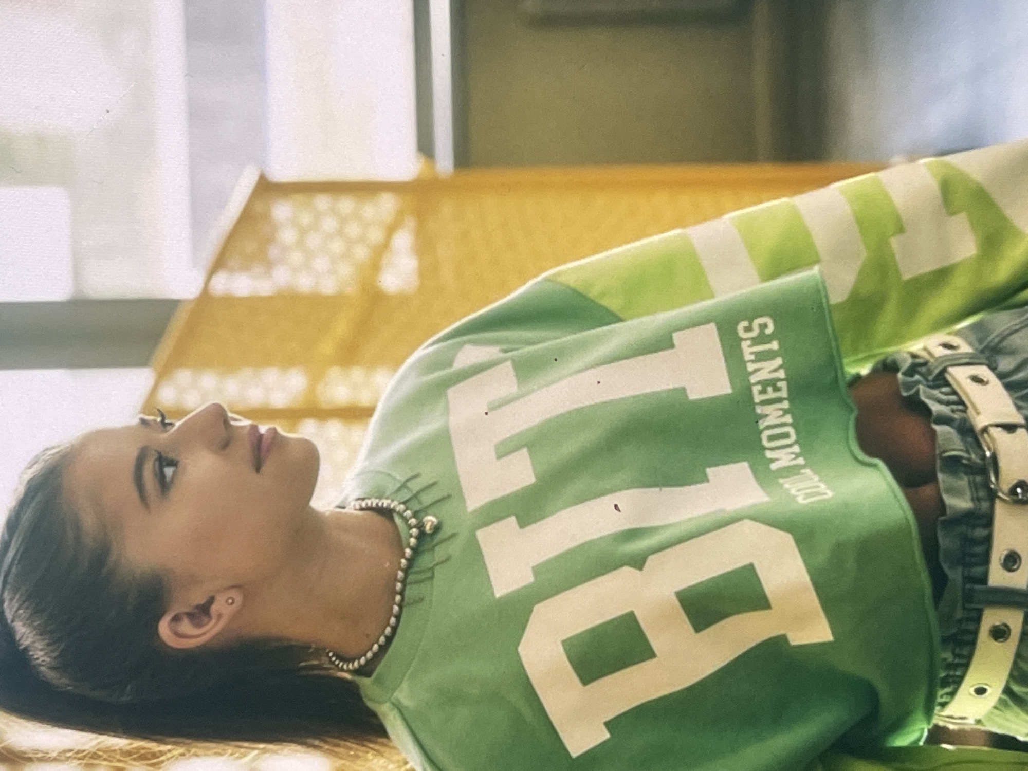 A young woman is captured in a side profile, lying on her back and gazing upwards. She has dark hair, a subtle ear piercing, and wears a silver beaded necklace. Her attire consists of a cropped hoodie or sweatshirt that transitions from light green to lime green. Prominently displayed on the front of the sweatshirt are large white letters "BLT," with additional vertical text "OOL MOMENTS" (likely "COOL MOMENTS") positioned below and to the right. She is also wearing blue jeans and a white belt with visible metal eyelets.
The woman's expression appears calm and serene, suggesting a moment of relaxation or contemplation. She is positioned on a textured surface, possibly a woven mat or piece of furniture with a distinct yellow-gold lattice pattern. The background is softly out of focus, indicating an indoor setting with subtle lighting and indistinct architectural elements. The image provides no specific time of day or weather cues, but implies a well-lit interior. The location context for this image is Poltava, Ukraine.
