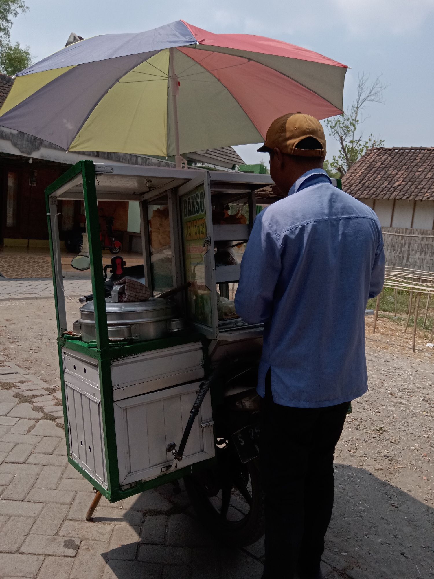 In Jumput, Indonesia, a man wearing a blue shirt and a brown cap stands behind a mobile food cart. The cart is equipped with an umbrella that has alternating panels of red, yellow, and gray. The cart itself is constructed with a green frame and appears to have glass panels, displaying food items within. A large metal pot is visible inside the cart. The ground is paved with cobblestones in the foreground, transitioning to a gravelly surface in the background. In the distance, there are buildings with tiled roofs and wooden fences. The sky is clear and bright.