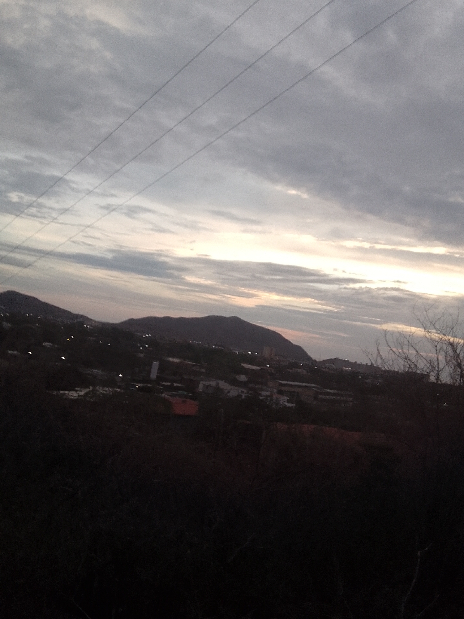 The image depicts an evening scene in Juan Griego, Venezuela. The sky is overcast with a pale sunset glow visible on the horizon. Power lines stretch across the upper left portion of the frame. A silhouette of a mountain range is visible in the middle distance, with faint lights suggesting a populated area at the base. The foreground is dominated by dark, undefined vegetation, likely shrubbery.
