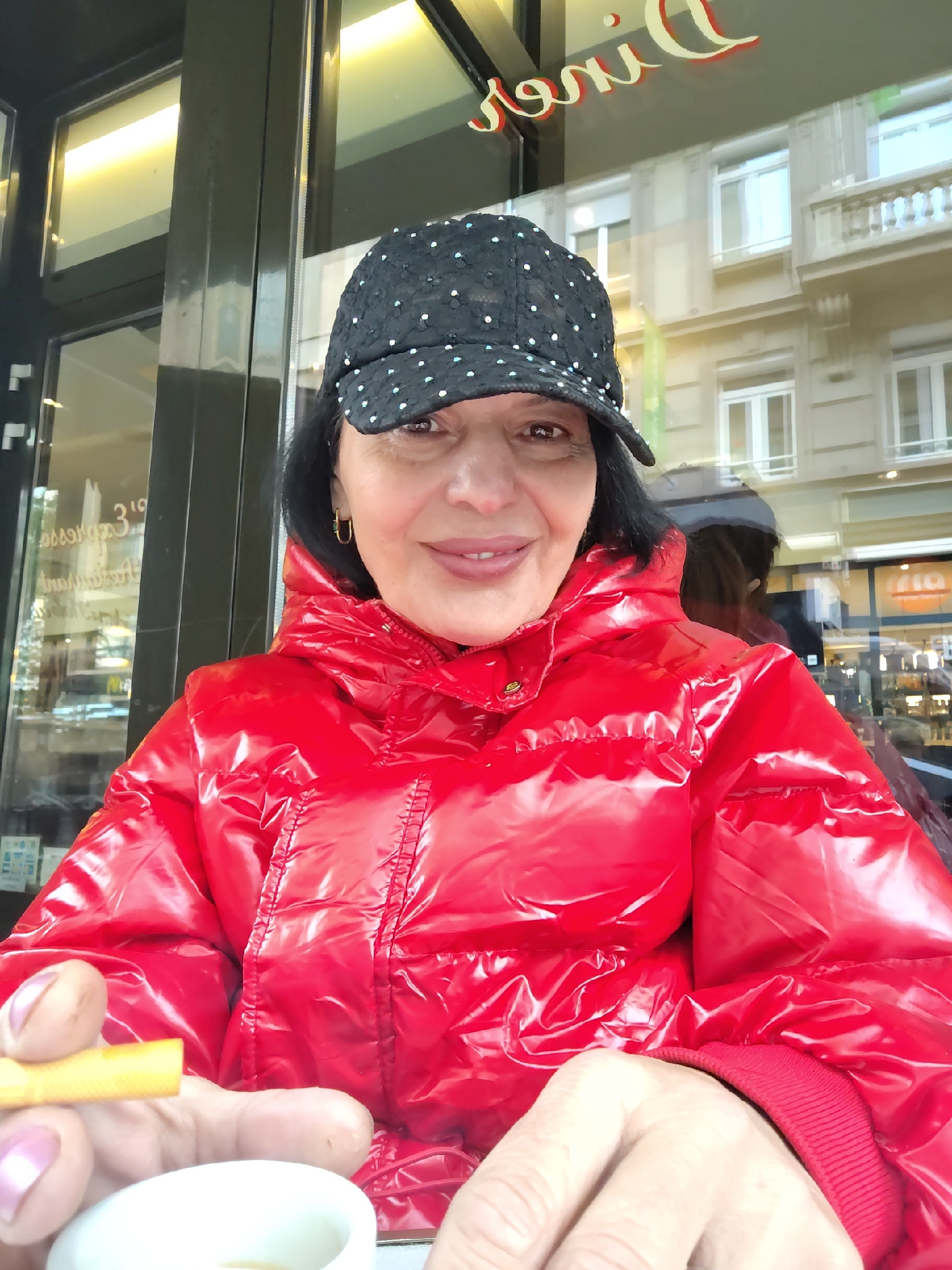The image shows a woman sitting at a table outside a café in Strasbourg, France. She is wearing a red, shiny puffer jacket and a black baseball cap with white polka dots. She is holding a yellow object (possibly a breadstick or small cigar) in one hand and there is a small white cup in front of her. The café appears to be called "Diner's" based on the reflection in the window. The architecture reflected in the windows indicates that the café is located in a relatively urban area with multi-story buildings across the street.