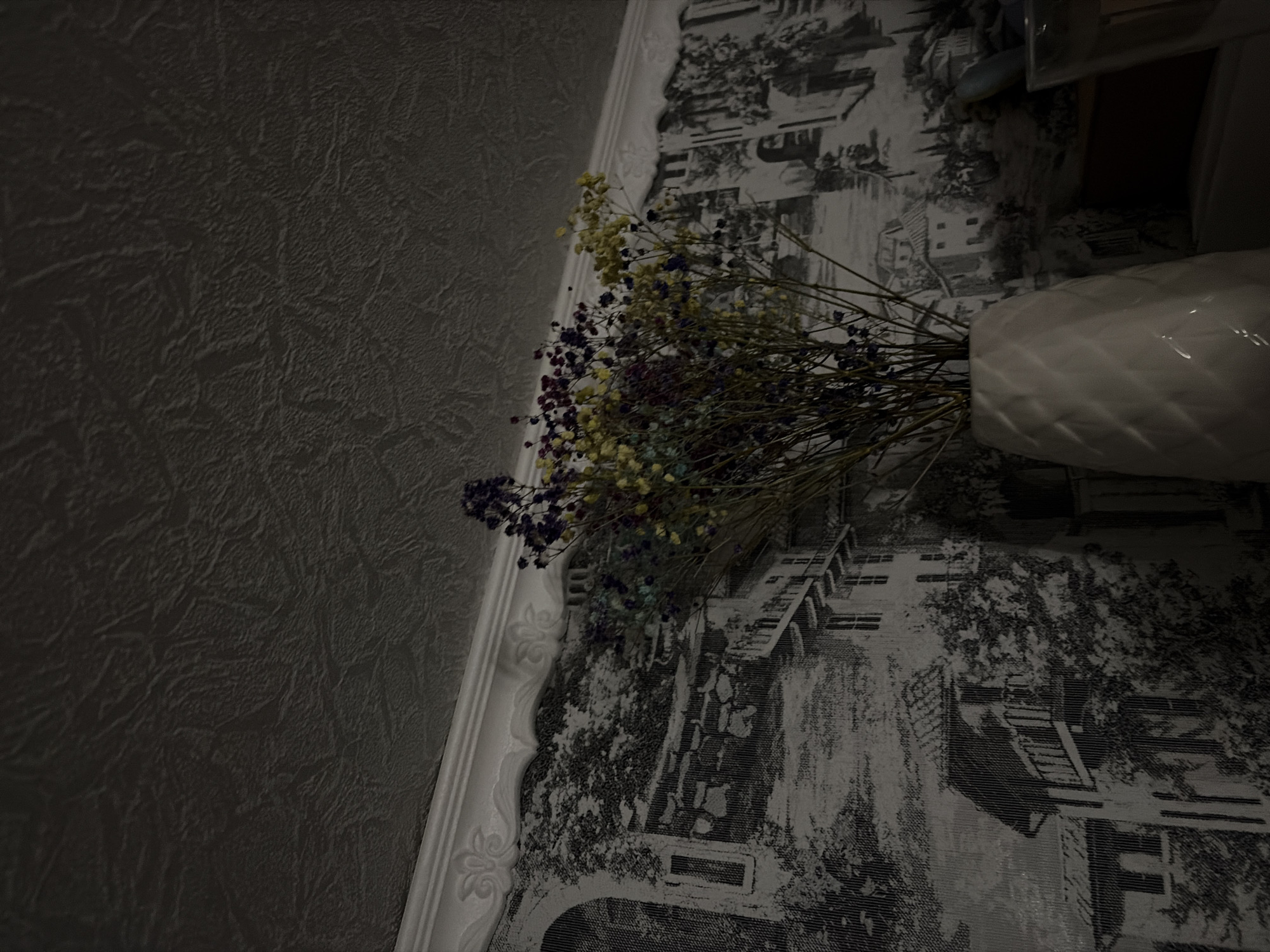 A dimly lit, tilted, close-up shot shows a white vase filled with a dried flower arrangement set against a textured gray wall and a black and white patterned wall. The flower arrangement is composed of small yellow, purple, and blue flowers. The vase has a ribbed texture. The patterned wall has a repeating design of what appears to be houses and trees. The wall is adorned with decorative moulding. There is no indication of the location being Новинки, Russia.
