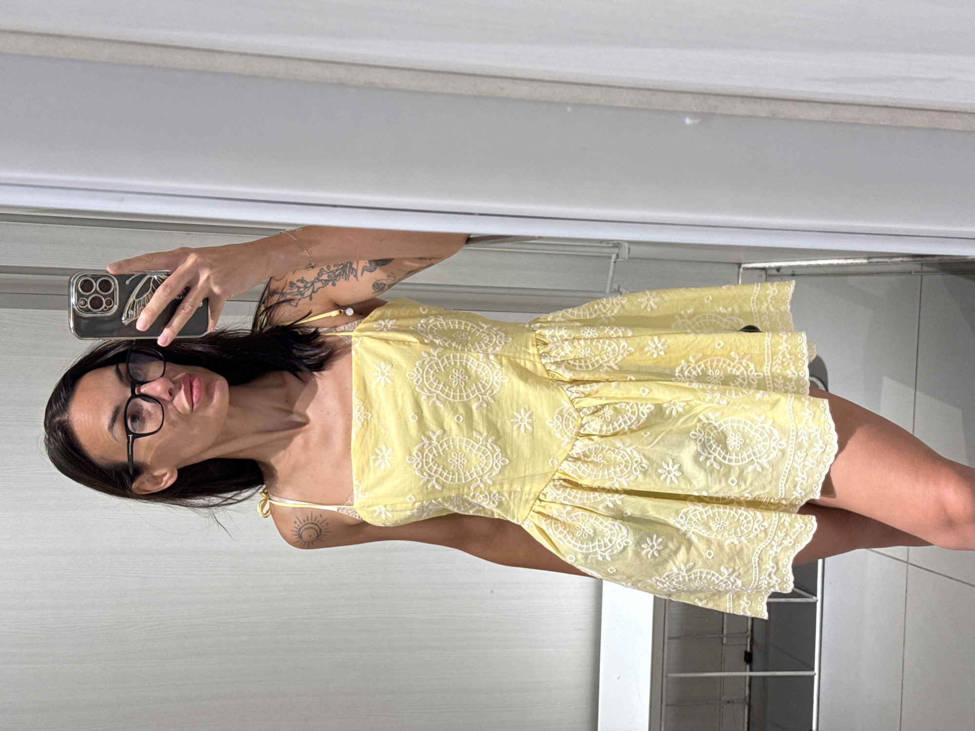 A woman with dark hair and glasses is taking a selfie in a mirror. She is wearing a yellow dress with white embroidery and holding a smartphone in her right hand. She has tattoos on her left arm and shoulder. The background appears to be a fitting room with white walls and a shelf. The image was taken in Pleven, Bulgaria.
