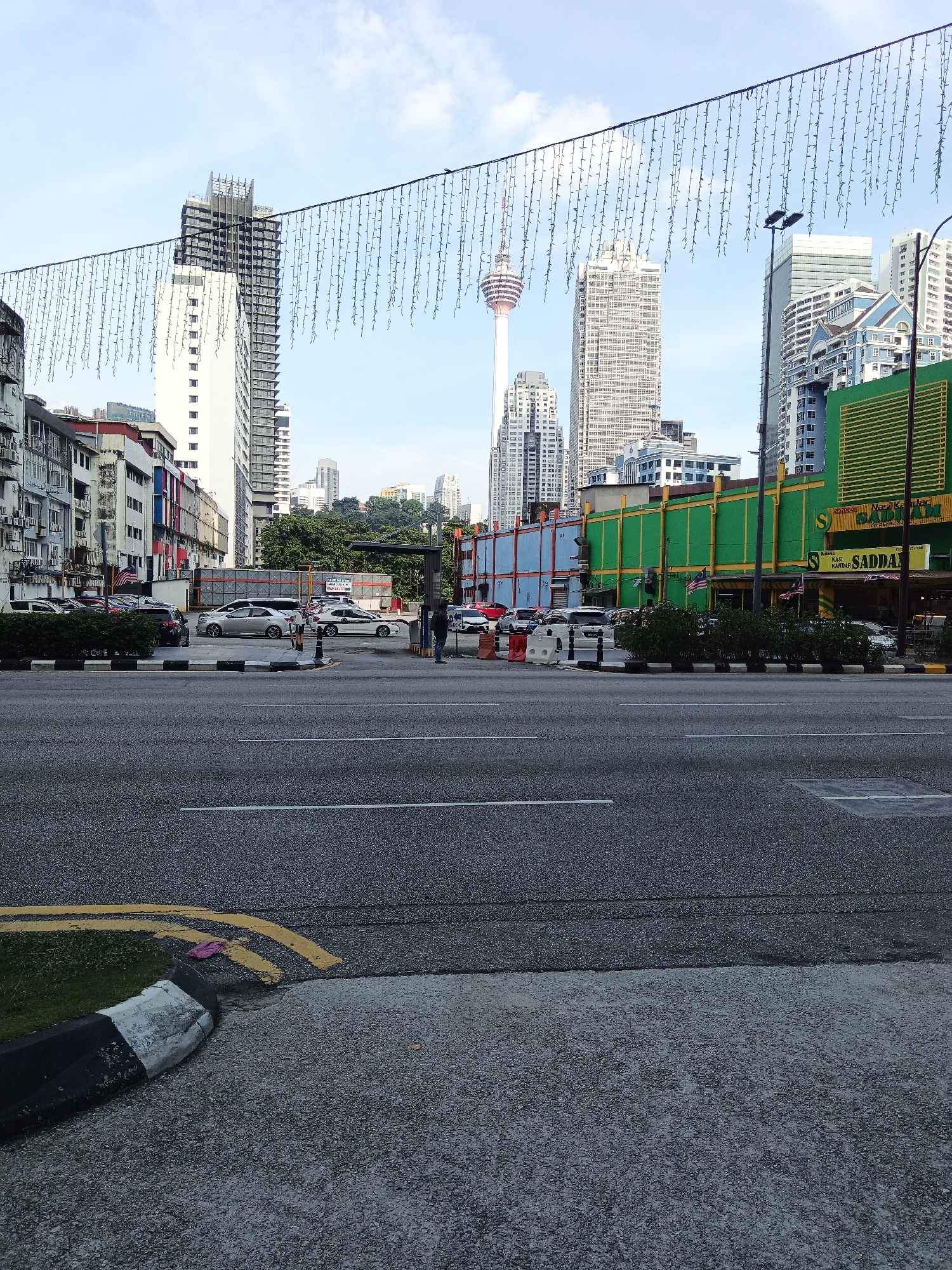 The image depicts a street scene in Kuala Lumpur, Malaysia, with the iconic Kuala Lumpur Tower prominently visible in the background. The foreground shows a paved road with yellow and white lane markings. To the right, a green building with yellow trim is visible, identified by signage as "Nasi Kandar SADDAM." Several cars are parked along the street. In the middle ground, a person in dark clothing stands near a bus stop shelter. Overhead, a string of decorative lights is suspended across the street. The sky is partly cloudy. The scene appears to be a commercial area with a mix of modern and older buildings.