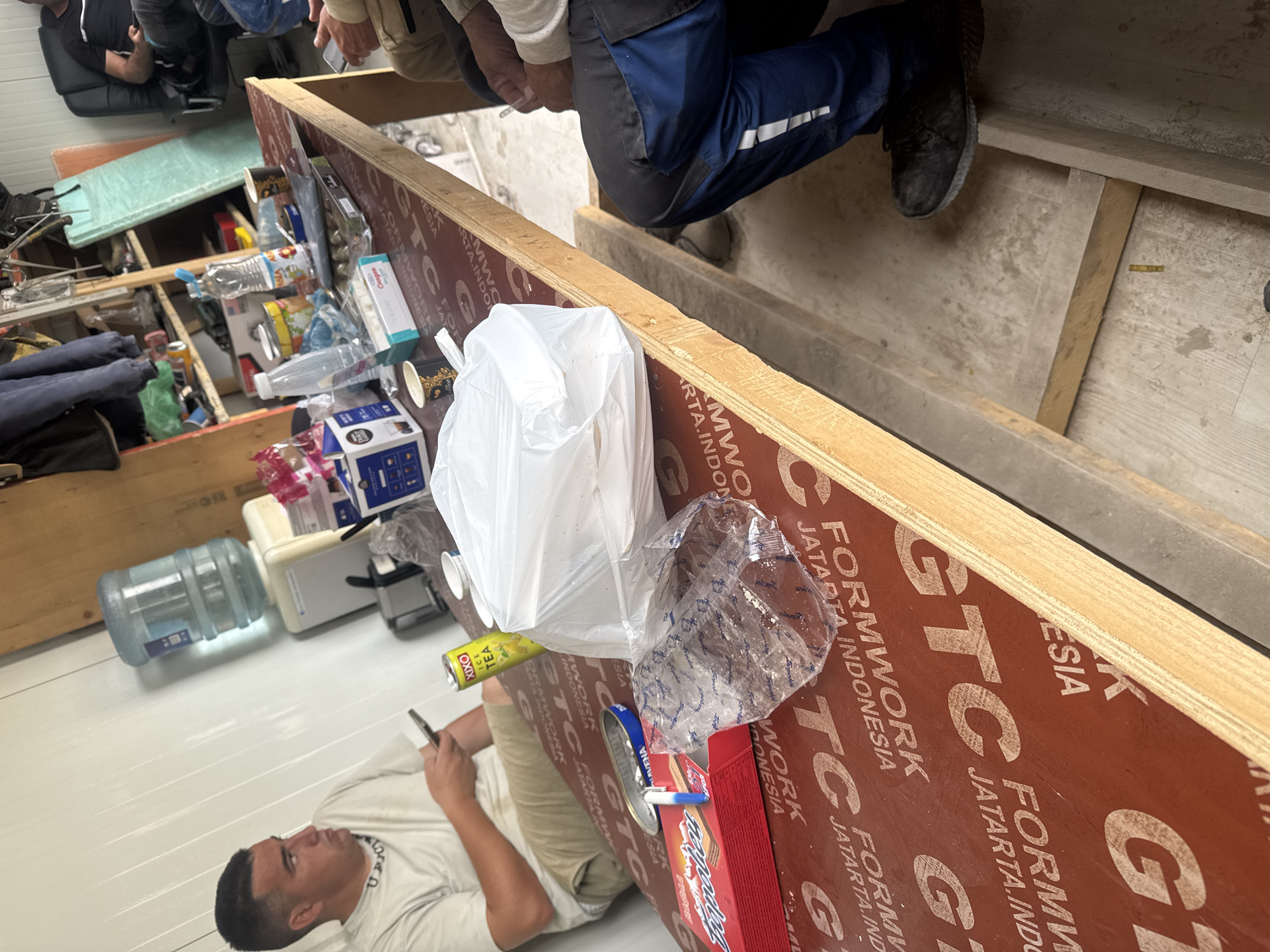 The image shows an indoor space with a chaotic and cluttered environment. In the foreground, there's a person lying down, looking at a phone. Nearby are discarded items, including a water bottle, a can labeled "XIXO Ice Tea," a box of wafers, and various plastic bags. The "Formwork GTC Jatarta Indonesia" is visible, suggesting the scene might be related to construction or remodeling. The lighting appears to be from fluorescent or LED fixtures, giving the space a sterile feel. The presence of multiple people and the scattered objects indicate a possible break area or temporary living space on a job site. The location of the image is not in Plovdiv, Bulgaria.