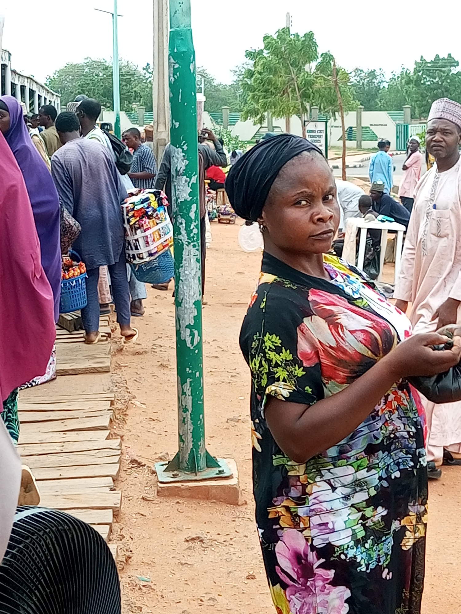 The image depicts a crowded outdoor scene in Gombe, Nigeria, likely a market or a transportation hub. In the foreground, a woman wearing a black headscarf and a floral print dress is the primary subject, looking towards the camera with a neutral expression. She is holding a small black plastic bag in her right hand. Behind her, a variety of people are visible, many in traditional clothing. To the left, a person in a purple hijab is partially visible. Several individuals are carrying bags, including a large white woven bag filled with colorful items, possibly snacks or small goods. A blue plastic basket is also visible. The ground is covered in sand, with a wooden walkway constructed from planks. A green metal pole, possibly a street light or part of a structure, stands prominently to the right of the frame. In the background, there are buildings, trees, and more people. The overall atmosphere suggests a busy, everyday scene in the city.
