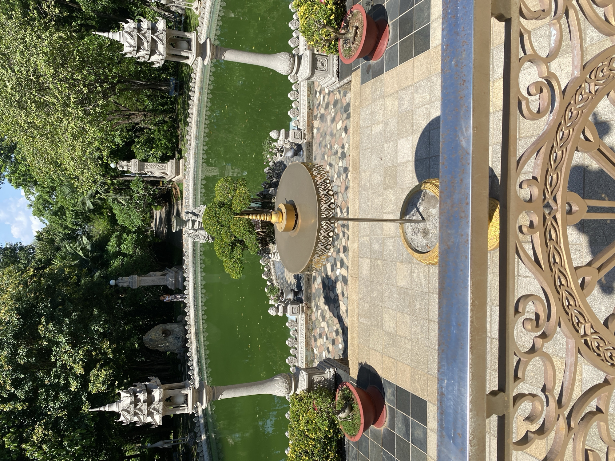 Overhead shot of a pool in Dĩ An, Vietnam, featuring green water and decorative statues along the edge. A fountain stands in the pool. Tile and potted plants decorate the surrounding area. A metal railing is in the foreground. Trees are visible in the background.