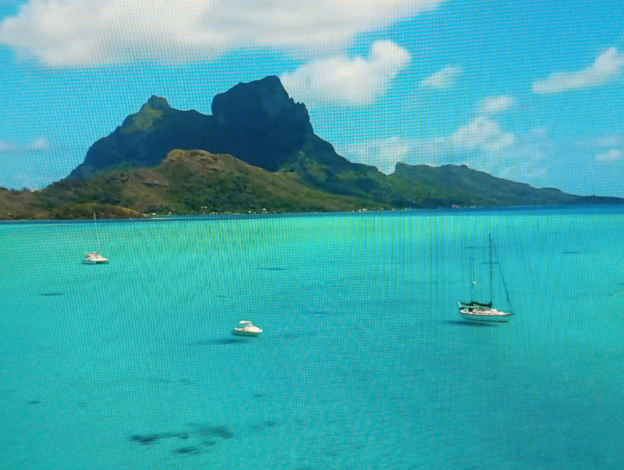 This is a photograph of a tropical island landscape. In the background, a large, mountainous island with lush green vegetation is visible under a bright blue sky with scattered white clouds. In the foreground, the crystal-clear turquoise water of the ocean is calm. Several boats are scattered across the water: a small white boat is positioned in the upper left quadrant, another small white boat is in the center, and a sailboat with a white hull and green trim is in the right quadrant. The water exhibits variations in color, with lighter patches indicating shallower areas. The image is a scenic view of a tropical paradise. There is no information in the image to suggest any connection to the city of Dubăsari town council, Moldova.