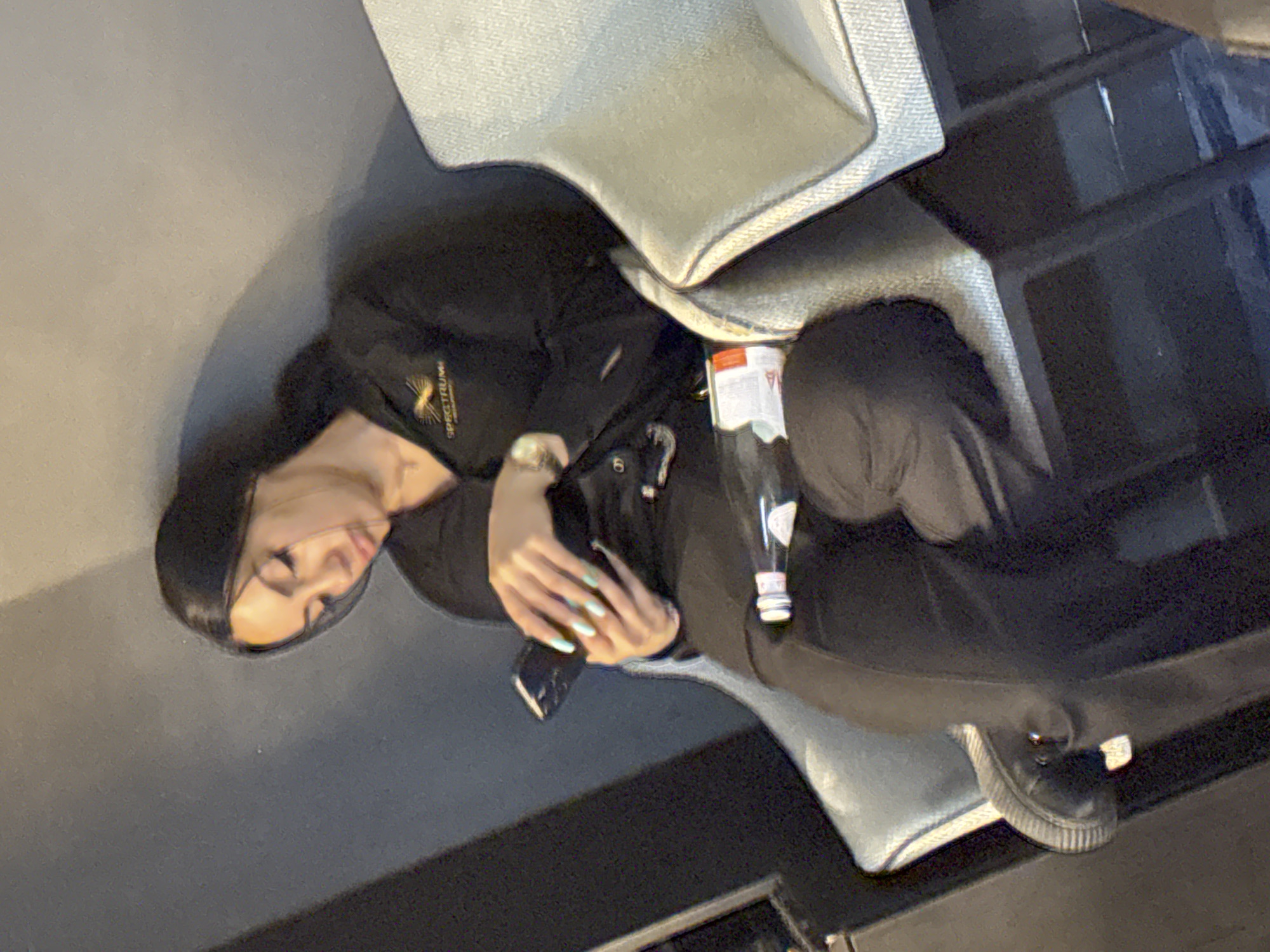 A woman with dark hair is sitting on a bench and looking at her phone. She is wearing a black shirt with a logo that says "SPECTRUM" and black pants. She has light blue nail polish on her fingernails. There is a small bottle on the bench next to her. The background is a gray wall and a light-colored chair. The image appears to have been taken indoors, possibly in a waiting area or lounge. There is no information in the image to indicate the location in Sofia, Bulgaria.