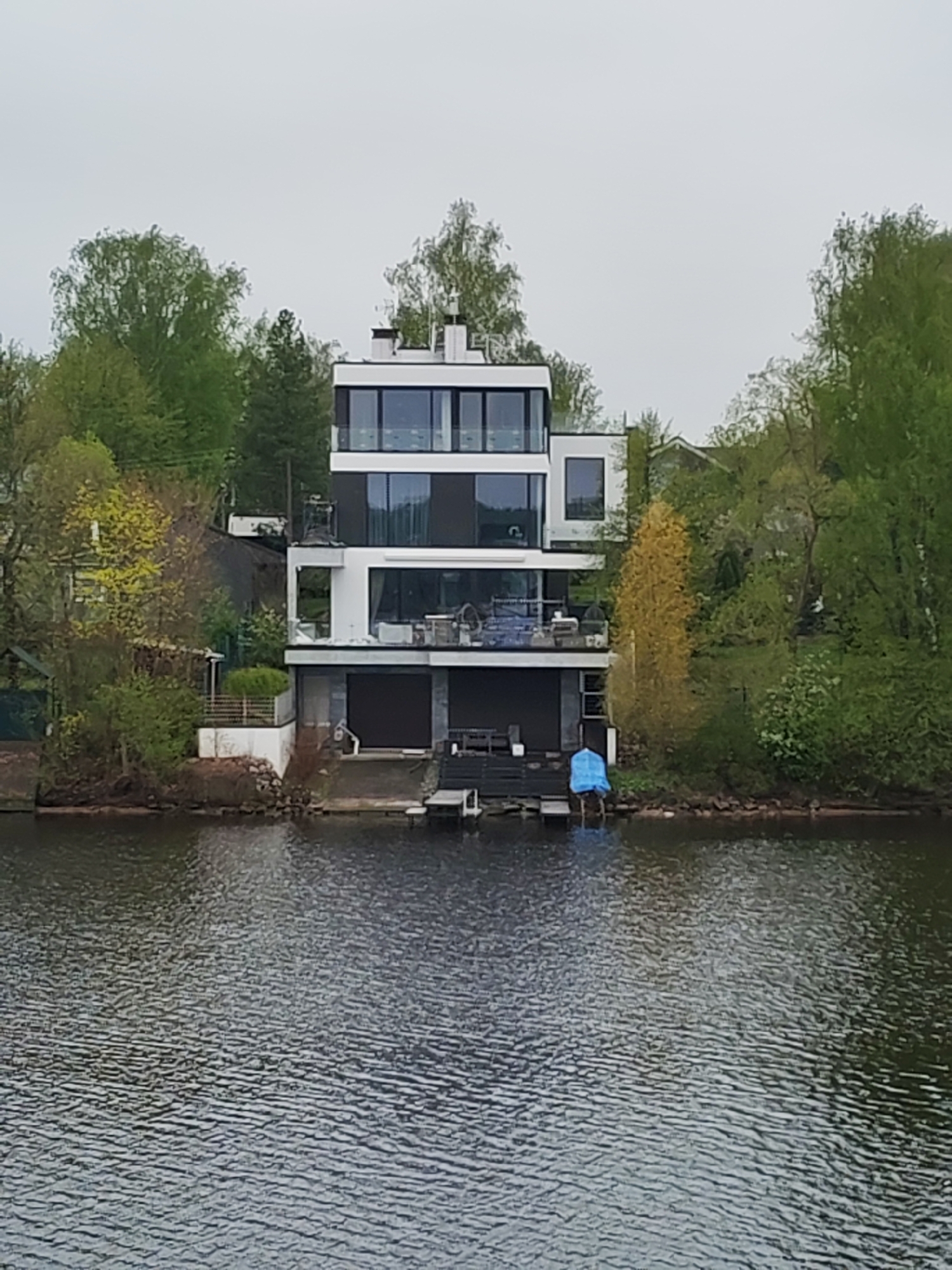 The image shows a modern, three-story house situated on the edge of a body of water. The house features a white facade with dark window frames and multiple balconies. The ground level appears to have closed shutters or garage doors. A small dock extends into the water in front of the house, with a blue cover visible near the water's edge. The house is surrounded by trees, with varying shades of green foliage, suggesting a lakeside or riverfront location. The sky is overcast. The image was taken in the city of Unknown, Unknown.
