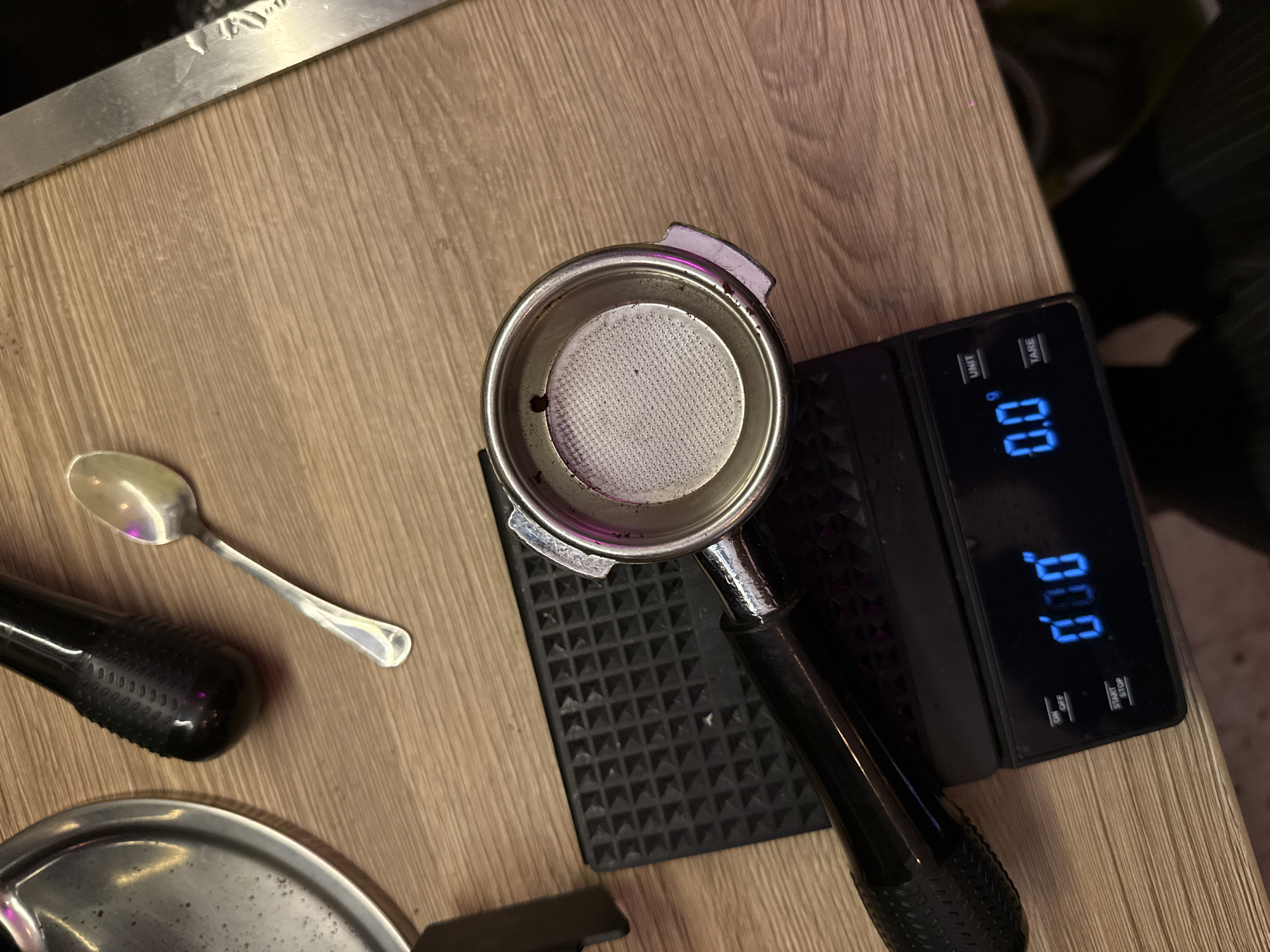 An overhead shot shows a wooden table with various items related to coffee preparation. In the center, a portafilter rests on a black tamping mat, which is placed on a digital scale displaying "0.0g". To the left of the portafilter is a shiny silver spoon. In the upper left corner, a portion of a metallic bowl is visible. Above the spoon and bowl, a black handle with textured grip is present. The right side of the image features a portion of a metallic object, possibly a knife. The background is a light-colored wooden surface. There is no indication of the city of Moscow, Russia.