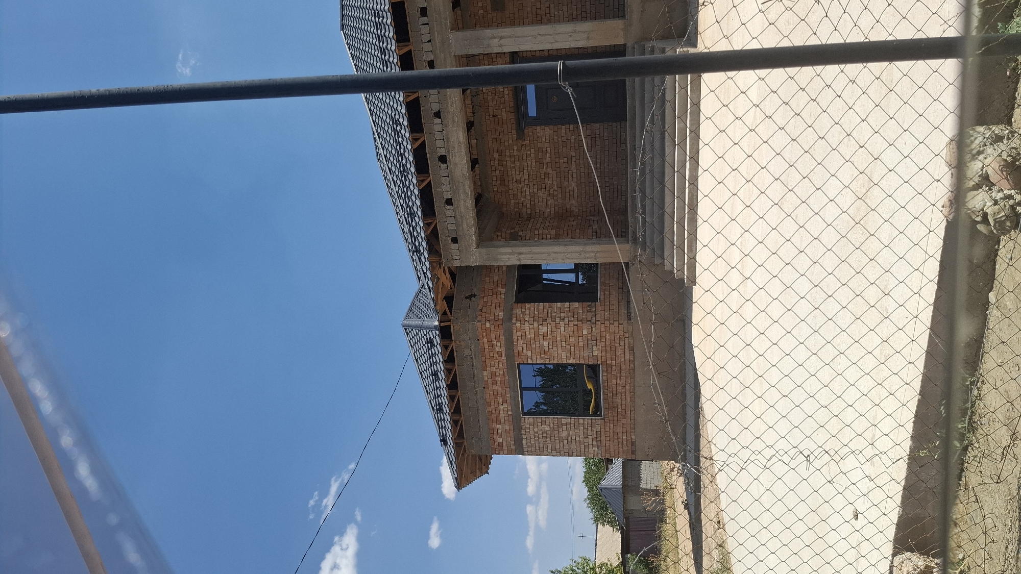 The image shows a brick house under construction in the city of Shubar asu, Kazakhstan. The house has a dark tiled roof, brick walls, and dark-framed windows. There are concrete stairs leading to the entrance. A chain-link fence is in the foreground, obscuring the lower part of the house and the ground. The sky is blue with some scattered clouds. The environment appears to be a residential area with other structures visible in the background.
