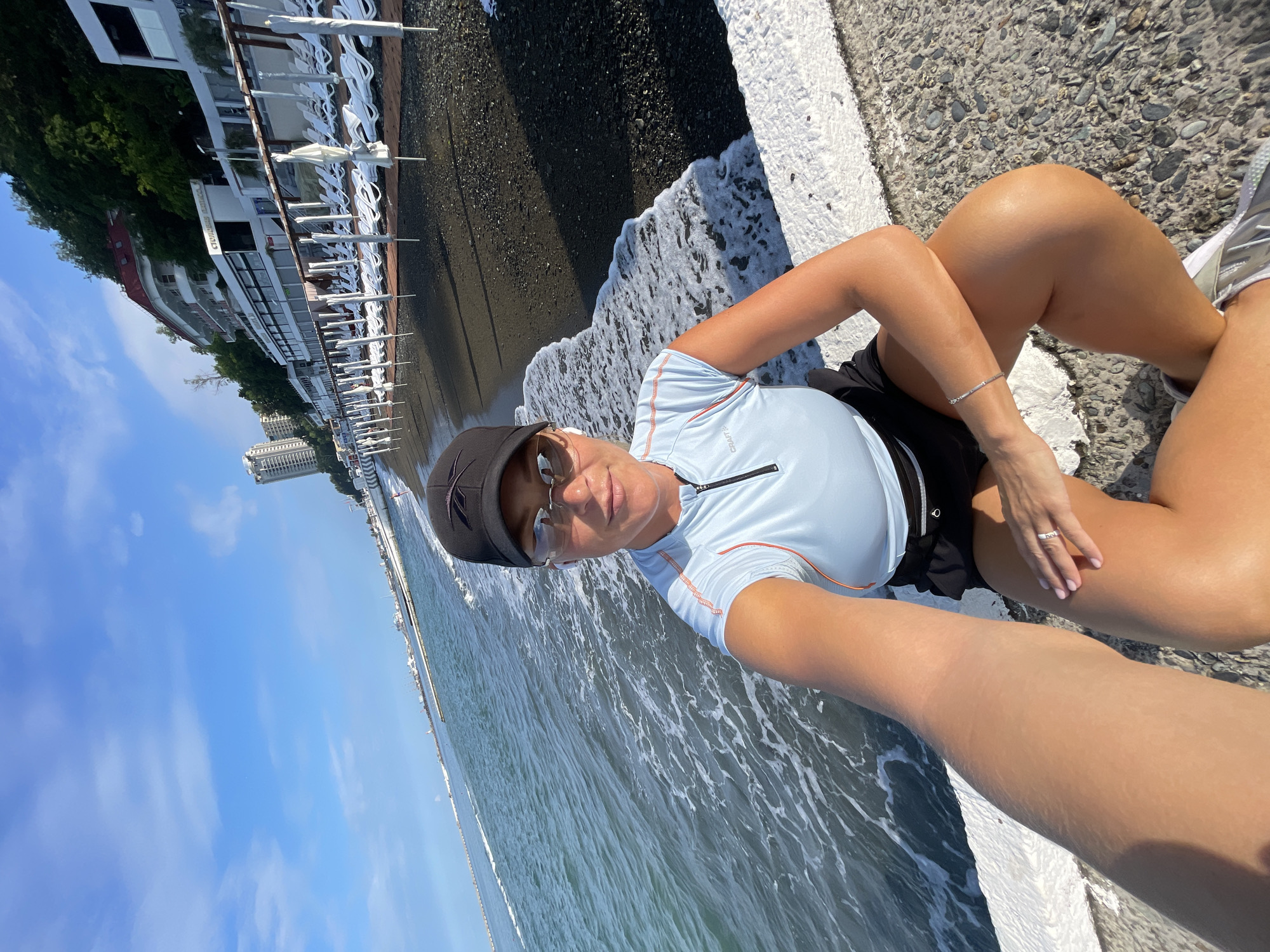 A woman wearing a light blue athletic top, black shorts, sunglasses, and a black baseball cap takes a selfie on a promenade next to the sea. The sea is on her left, with waves lapping against a white concrete barrier. Behind her, the promenade continues, lined with rows of white lounge chairs and umbrellas, and a multi-story hotel building sits on a hill overlooking the beach. In the distance, a tall, modern building is visible. The beach itself is dark and pebbly. The sky is bright blue with scattered white clouds, indicating a sunny day. The location is identified as a beach in Sochi, Russia.