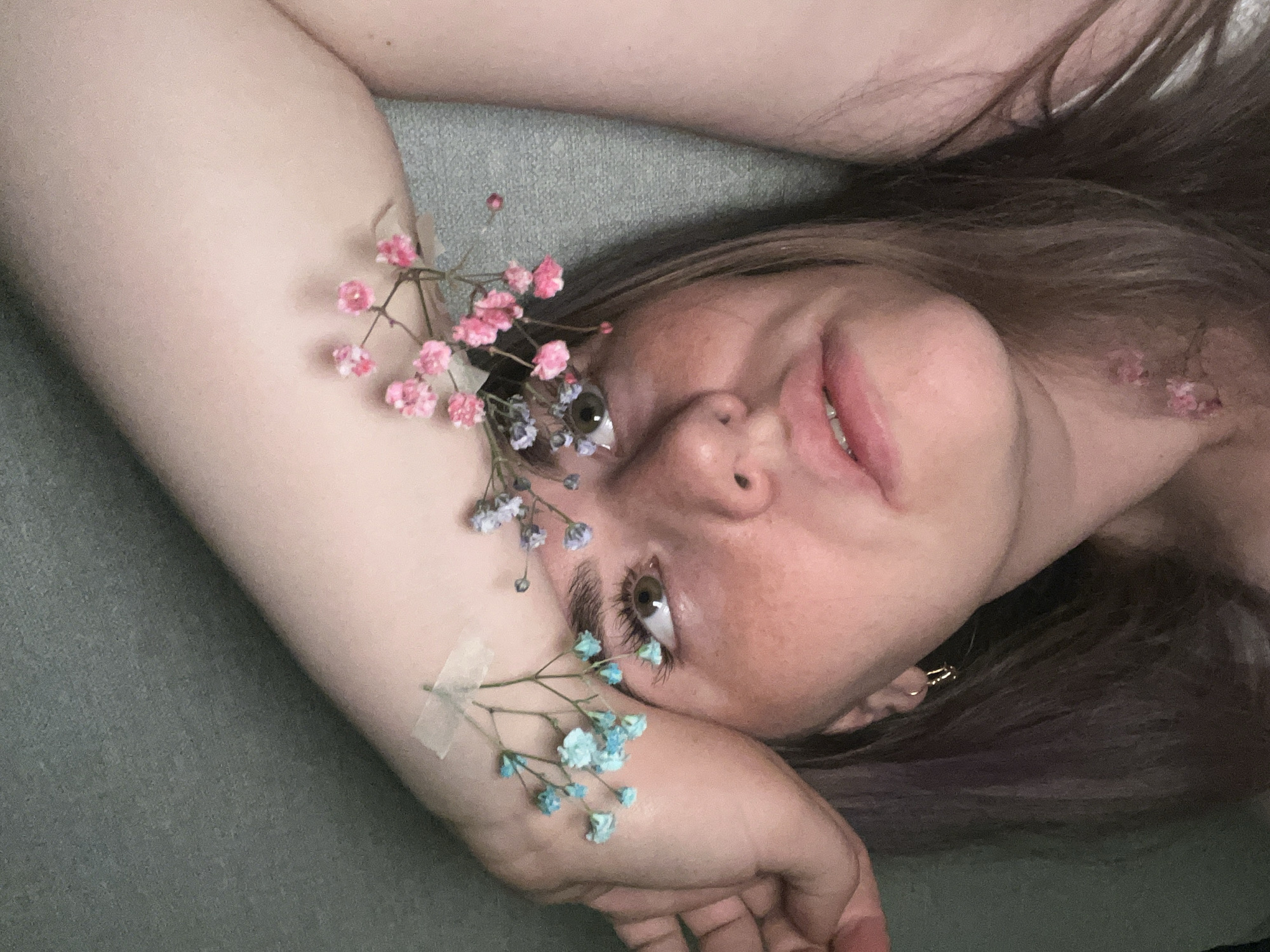 A woman lies on a grey-green surface, possibly fabric, with her head tilted back and to the right. Her brown hair is spread around her head. Pink and blue baby's breath flowers are arranged around her face and arm, some taped to her skin. She is looking up and to the left. No specific details indicate the location is Chaikovsky, Russia.