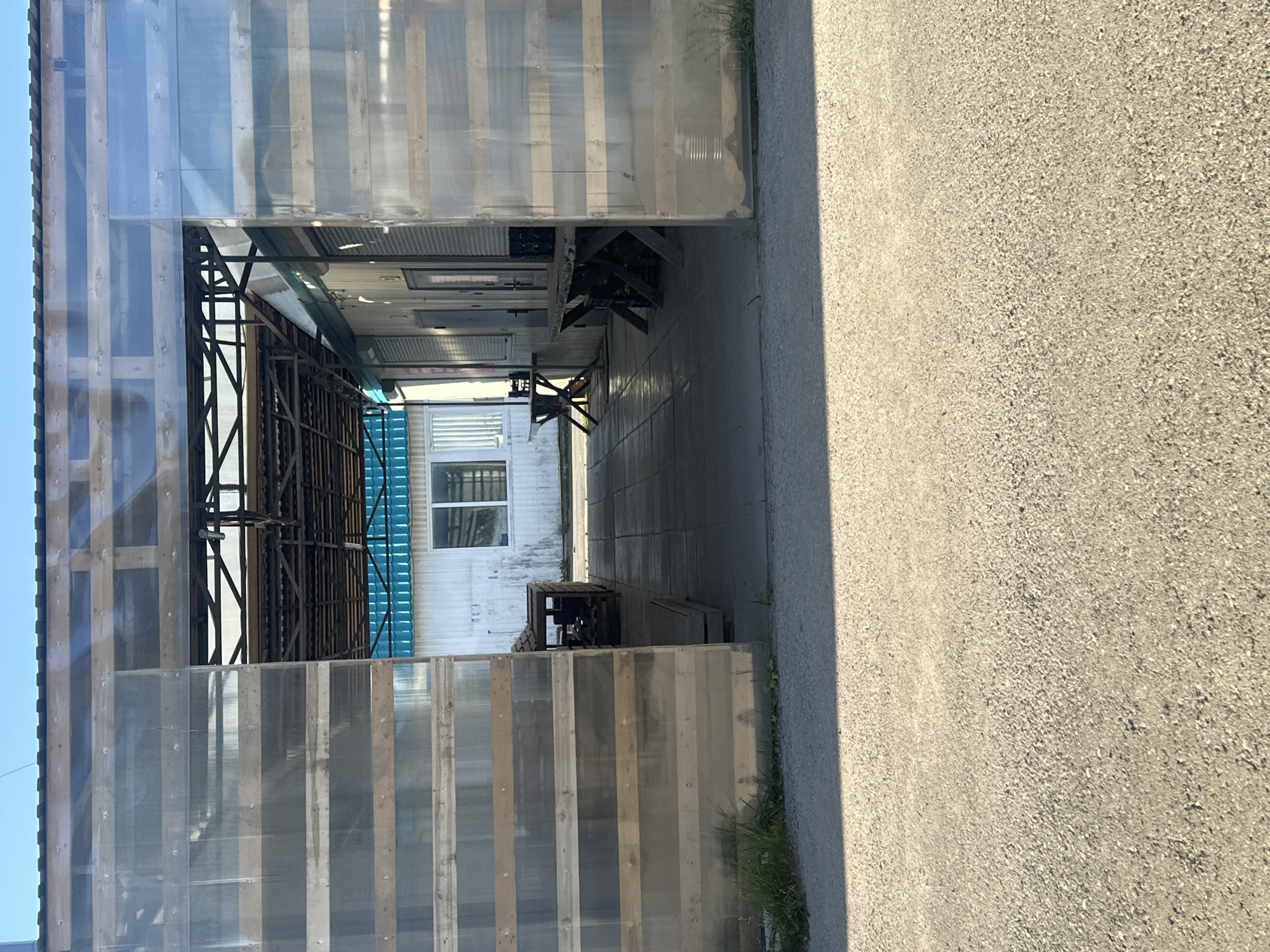 The image shows an outdoor area in Plesetsk, Russia, with a covered walkway or structure. The walkway has walls made of corrugated plastic or similar material over wooden planks. Inside the covered area, there are two tables with chairs, possibly for outdoor dining or seating. In the background, a white building with a blue roof and windows is visible. The ground is paved with square tiles, and a gravel or asphalt road is in the foreground. The scene appears to be a public space, possibly part of a market or a cafe. There are no people visible in the image.