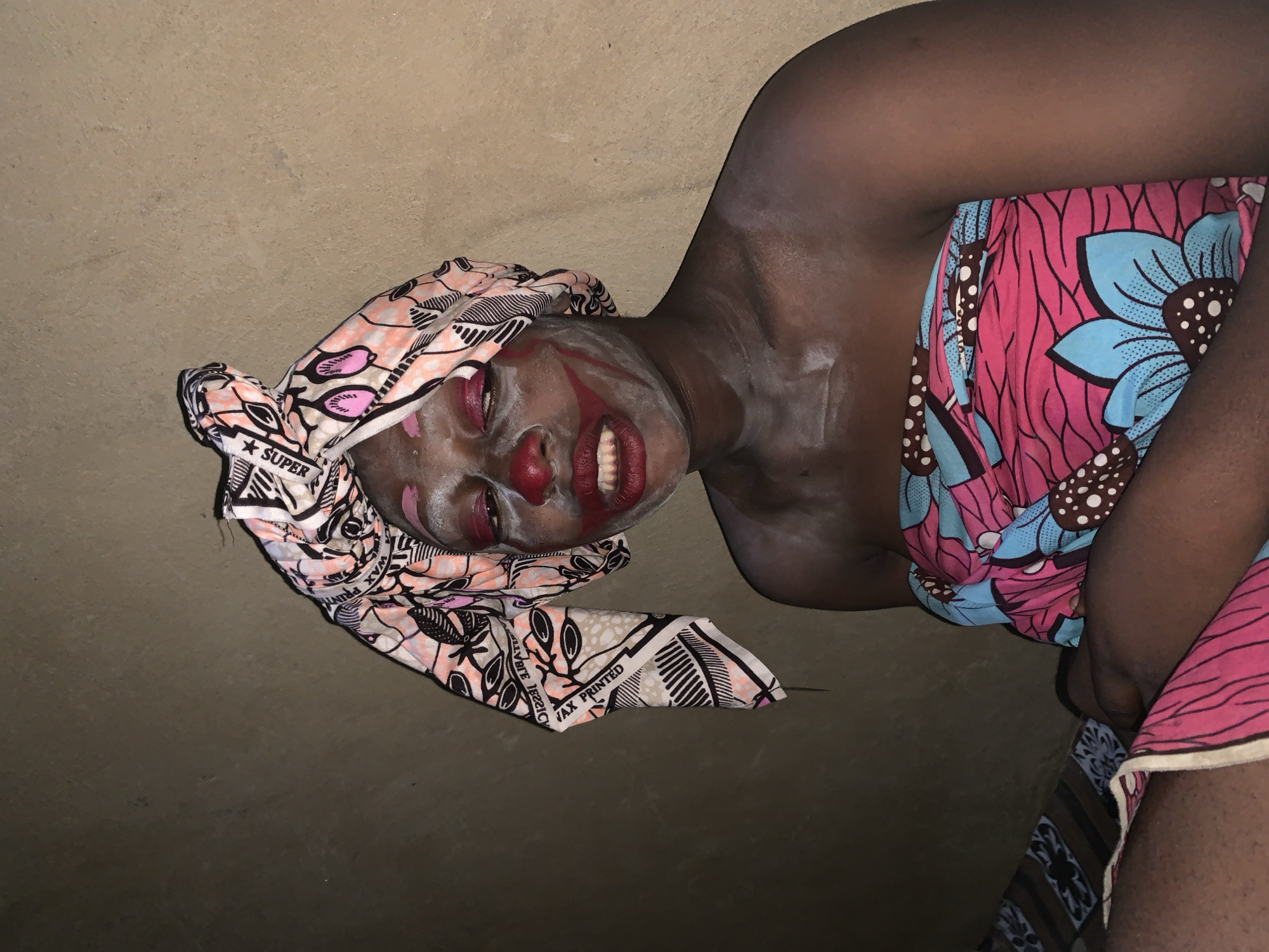 In Agban, Nigeria, a person with clown-like makeup, including a red nose and painted red lips and cheeks, poses in front of a textured, beige wall. They are wearing a colorful headscarf with a black and white patterned trim and a strapless, floral-print garment in shades of pink and blue. The person's skin is dusted with white powder, particularly on their chest and shoulders. A patterned textile, possibly a rug or blanket, is visible on the left side of the frame. The lighting suggests an indoor setting, possibly at night, with a single light source illuminating the subject.