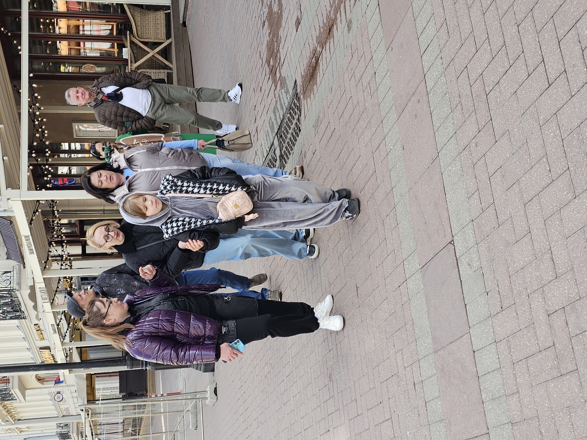 A photograph shows a group of six individuals standing on a brick-paved pedestrian walkway outside an Italian restaurant called "Dolccini" in the city of Moscow, Russia. The restaurant has a visible awning and outdoor seating. The individuals are lined up in a row, appearing to be posing for the photograph. They are dressed in casual clothing appropriate for an outdoor setting. The ground is paved with a mix of red and grey bricks in a herringbone pattern.
