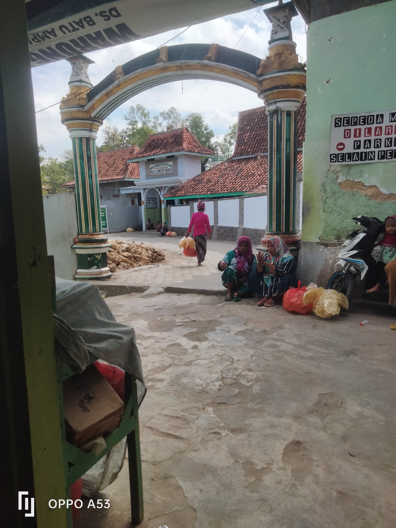 The image captures an outdoor scene in what appears to be a village or suburban area in Badung, Indonesia, possibly near a place of worship or community center, suggested by the signage and architectural style. The foreground is dominated by an arched entryway with decorative pillars, painted green and gold. To the right of the arch, a light green wall is visible, bearing a sign with black lettering on white backgrounds that reads "SEPEDA MOTOR DILARANG PARKIR SELAIN PETUGAS." This translates to "MOTORCYCLES FORBIDDEN TO PARK EXCEPT FOR OFFICERS."
In the mid-ground, a woman in a pink headscarf and dress is walking away from the camera, carrying two large plastic bags filled with goods. She is walking on a paved pathway. To her left, near a pile of what appears to be dried straw or vegetation, are two women sitting on the ground. Both are wearing headscarves and traditional Indonesian clothing. One is looking towards the camera, while the other is gesturing with her hands. Red and yellow plastic bags are placed on the ground near them.
Further back, beyond the arch and pathway, is a building with red-tiled roofs, possibly a mosque or a community building. A sign on this building reads "SELAMAT DATANG" (Welcome) and then some text in Indonesian that is partially obscured, but appears to indicate the location "BATU AMPAR BARAT."
To the far right, a white and blue motorcycle is parked, with a young child sitting on it, facing the camera. Another person's legs are visible on the right edge of the frame.
The overall environment appears somewhat dry and dusty, with cracked pavement in some areas. The lighting suggests it is daytime with a partly cloudy sky. In the immediate foreground on the left, a green wooden structure is partially visible, draped with a grey cloth, and a cardboard box is placed on it. The OPPO A53 logo is present in the bottom left corner of the image.