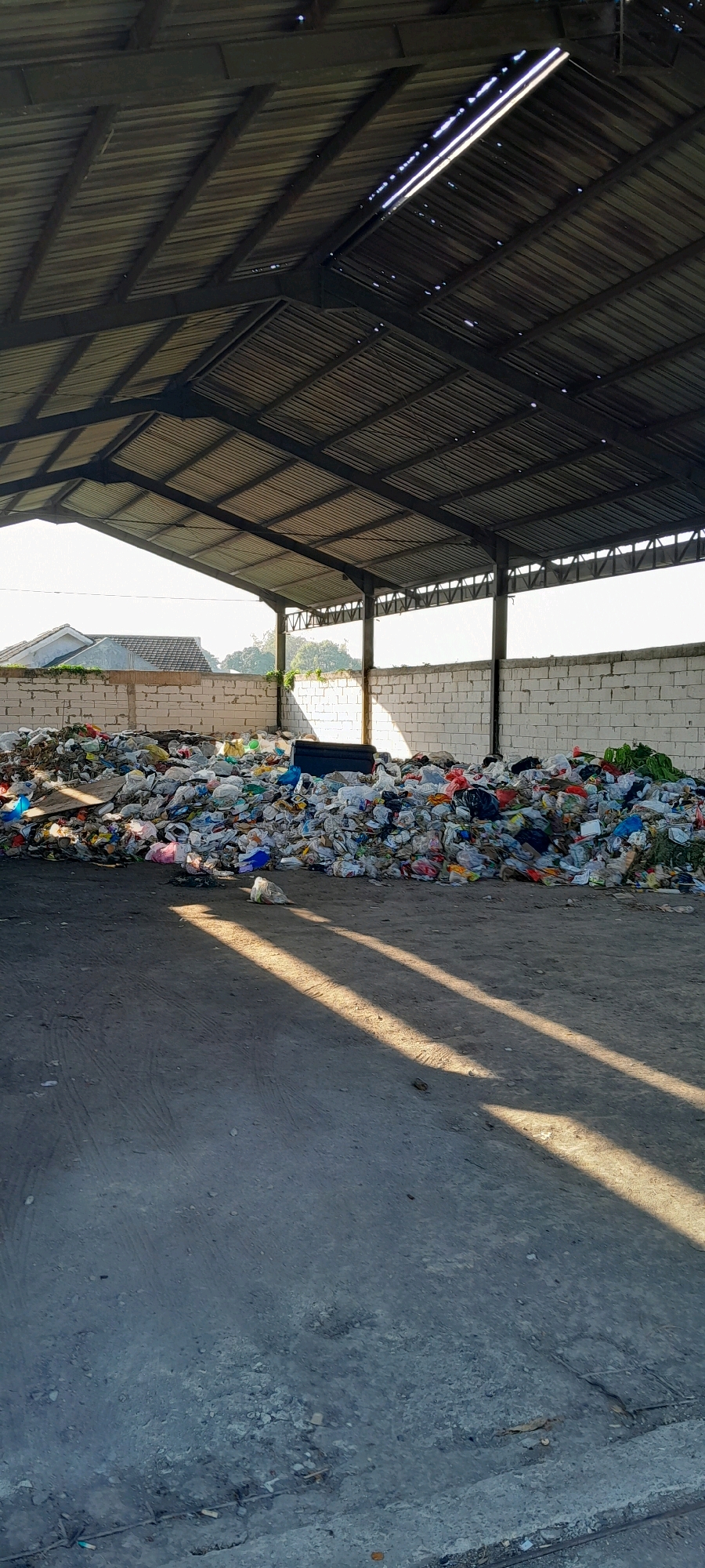 The image shows a large pile of mixed waste, predominantly plastic bags, bottles, and other refuse, accumulated inside a covered structure. The structure has a corrugated metal roof supported by dark metal beams and pillars, with open sides allowing natural light to enter. The waste is piled high against a white brick wall that forms the rear of the structure. Outside the structure, visible through the open front, are residential buildings with tiled roofs and some greenery, suggesting an urban or semi-urban environment. The ground in front of the waste pile is a rough, paved surface, with distinct sunlight and shadow patterns cast by the roof structure. The overall scene depicts a waste accumulation or sorting area. This location is identified as being in Bendungan, Indonesia.
