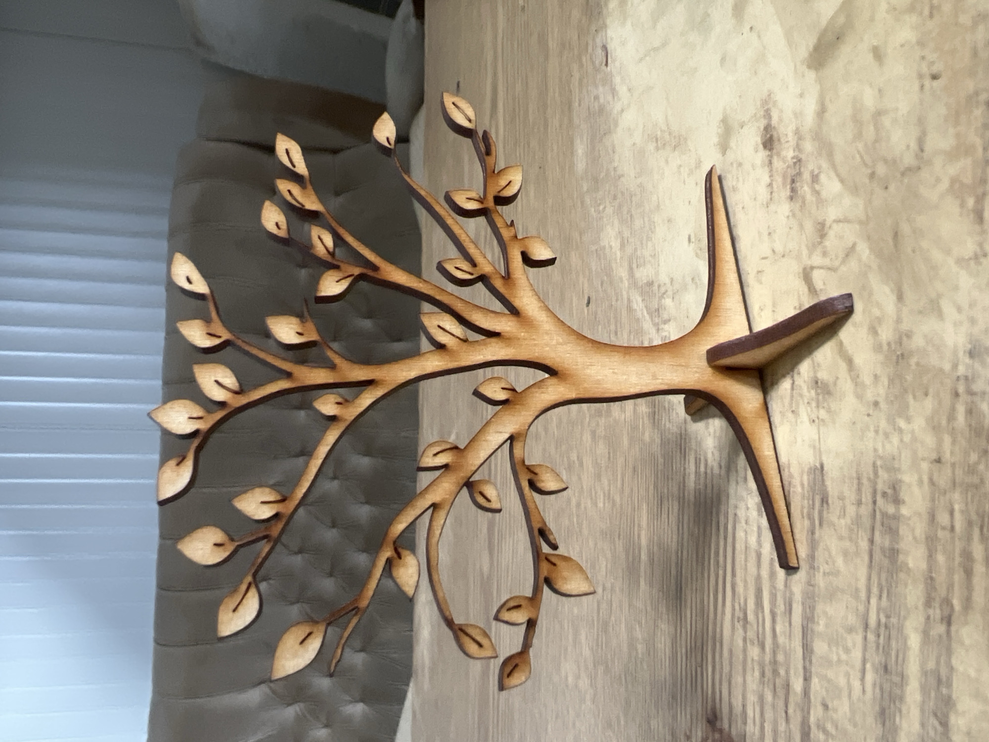 A laser-cut wooden tree branch stands on a light-brown wooden surface. The background consists of a grey upholstered object and a vertical-striped covering. The location is not discernible from the image and cannot be specifically identified as Burgas, Bulgaria.