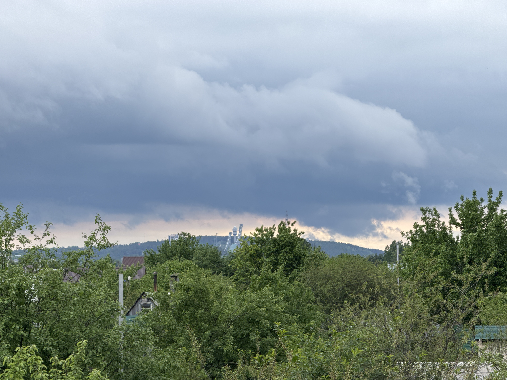 The image shows a view from within Chaikovsky, Russia. Green trees and foliage fill the lower portion of the frame, obscuring much of the foreground. Behind the trees, there's a glimpse of rooftops of small buildings or houses. In the midground, a hill or mountain range is visible, with a clear view of the "Snezhinka" ski jumping complex, a prominent landmark in Chaikovsky. The sky is overcast with heavy, gray clouds dominating the upper portion of the image.