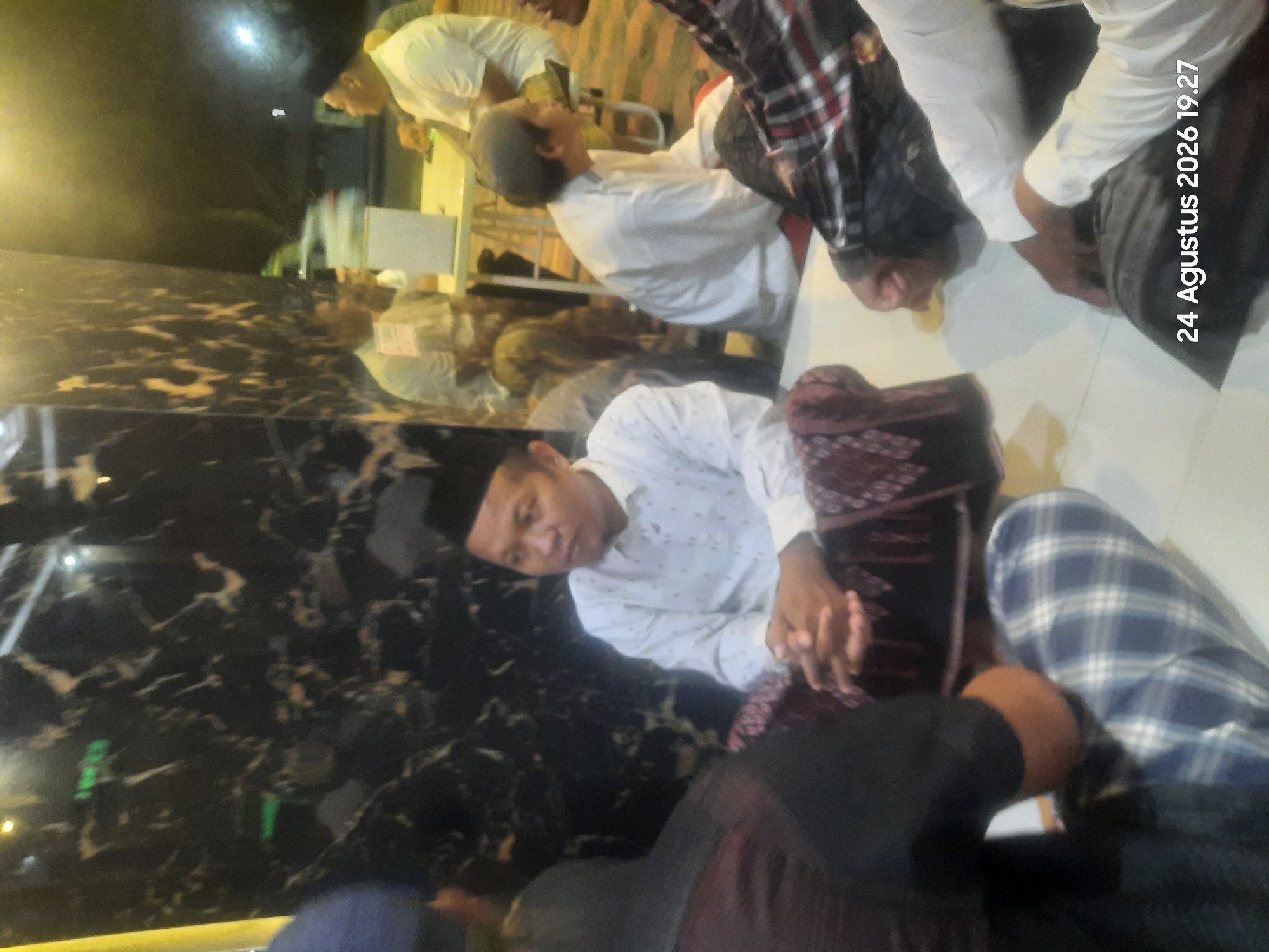 The image depicts a group of men, some wearing traditional Islamic attire such as peci caps and sarongs, gathered indoors. One man in the foreground, wearing a white patterned shirt and a peci, is seated with his hands clasped. To his left, another man is partially visible from the back, wearing a black shirt. Further back, several other men are seen seated, with one in a white shirt and a grey cap. The setting appears to be a public indoor space, possibly a mosque or a communal hall, with a dark, reflective wall in the background that shows faint reflections of lights and people. A white table with some objects on it is visible in the middle ground. The overall lighting suggests it is evening. The lower right corner of the image contains a timestamp: "24 Agustus 2026 19.27". The reference to Sukokerto, Indonesia is an external piece of information not directly verifiable from the image itself.