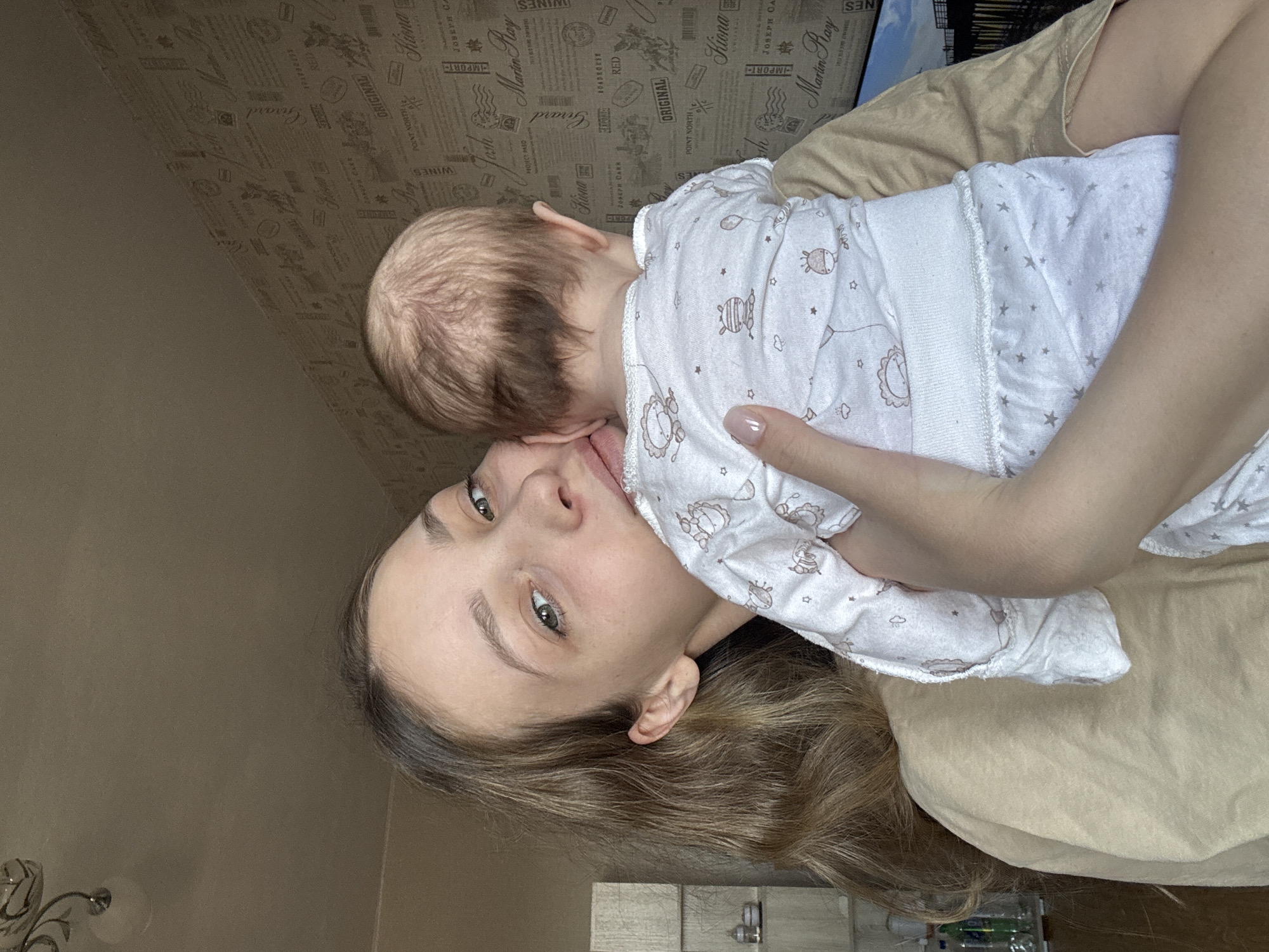 A woman with light skin, light brown hair, and green eyes is holding a baby with a fair complexion and light hair. The woman is looking at the camera with a slight smile. The baby is lying in her arms, facing away from the camera. The baby is wearing a white onesie with a pattern of stars and cartoon animals. The woman is wearing a beige shirt. The background is a patterned wallpaper. The image appears to be taken in Chaikovsky, Russia. There is a chandelier on the left side of the frame and a window on the right side. In the bottom of the image is a portion of a table with items on top.