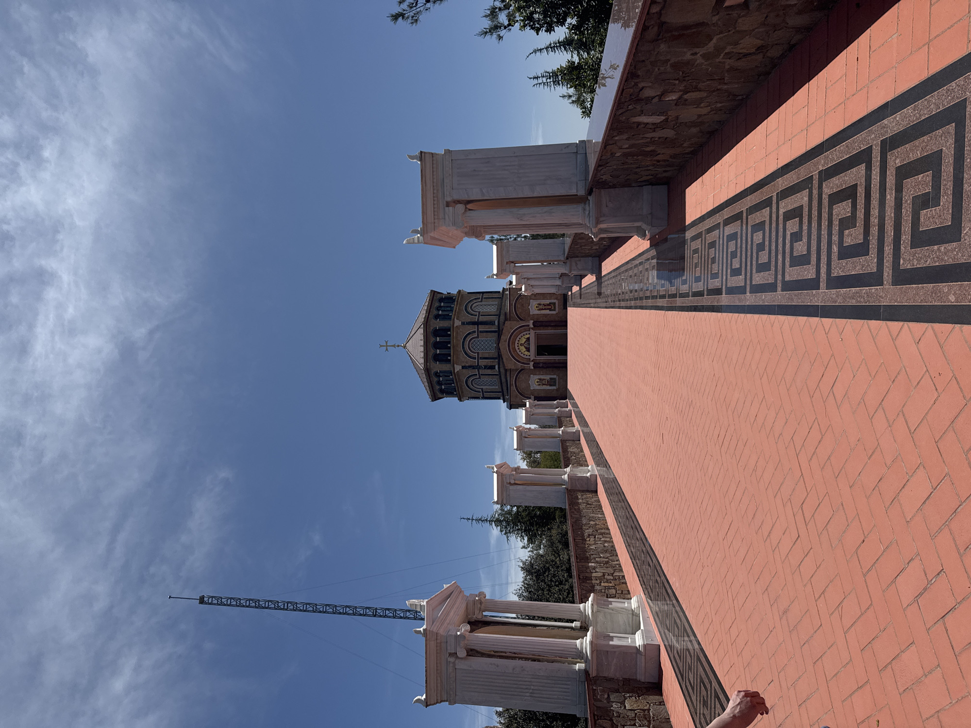 The image shows the Church of Panagia tou Moutoulla in Tsakistra, Cyprus, from a low-angle perspective. The walkway leading to the church is paved with red bricks, and lined with a decorative band featuring a Greek key pattern. Along the walkway stand marble columns. In the distance is the ornate church with a dome and cross. The sky is blue with scattered clouds. A crane is visible in the background to the left of the church. A human hand is visible on the lower right corner.