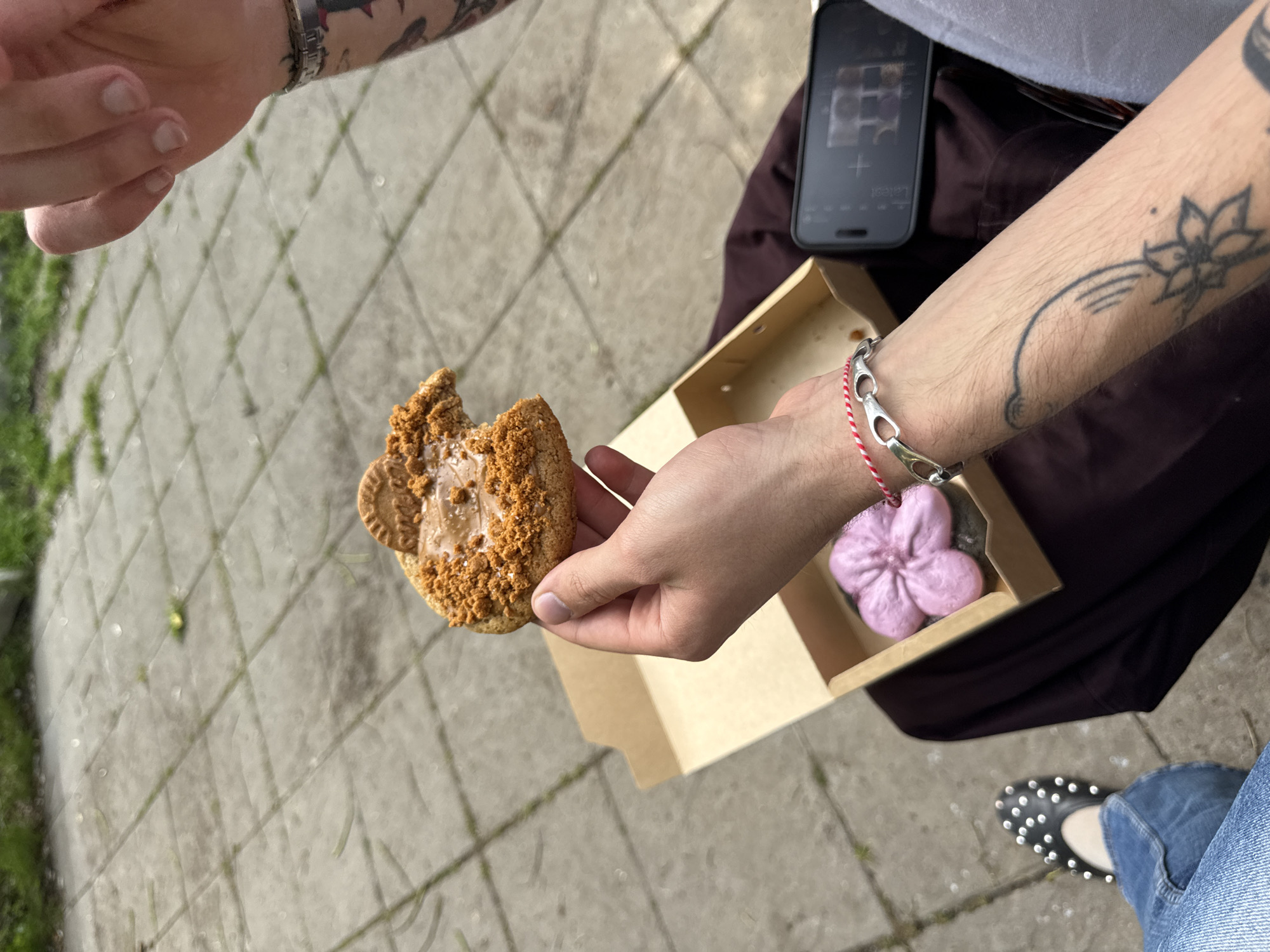 The image captures an outdoor, high-angle view of two individuals interacting on a paved surface in Sofia, Bulgaria. The primary subject, seated or resting, has their right arm extended, holding a large, partially eaten cookie topped with a spread and cookie crumbs, featuring a small biscuit on its surface. This person's arm displays tattoos, including a prominent flower design, and wears a silver chain bracelet along with a red and white string bracelet.
On their lap, a small, light brown cardboard box contains a distinct pink, flower-shaped pastry or dessert. Beside the box, a black smartphone rests, displaying "16:21" and what appears to be a photo gallery or camera interface. The person is wearing dark clothing, blue jeans, and black studded flat shoes. A second person's hand is visible in the upper left corner, reaching into the frame, suggesting an interaction with the cookie or the person holding it. The setting is an outdoor area with square concrete pavers, with glimpses of green grass or weeds along the edges, under bright daylight conditions.