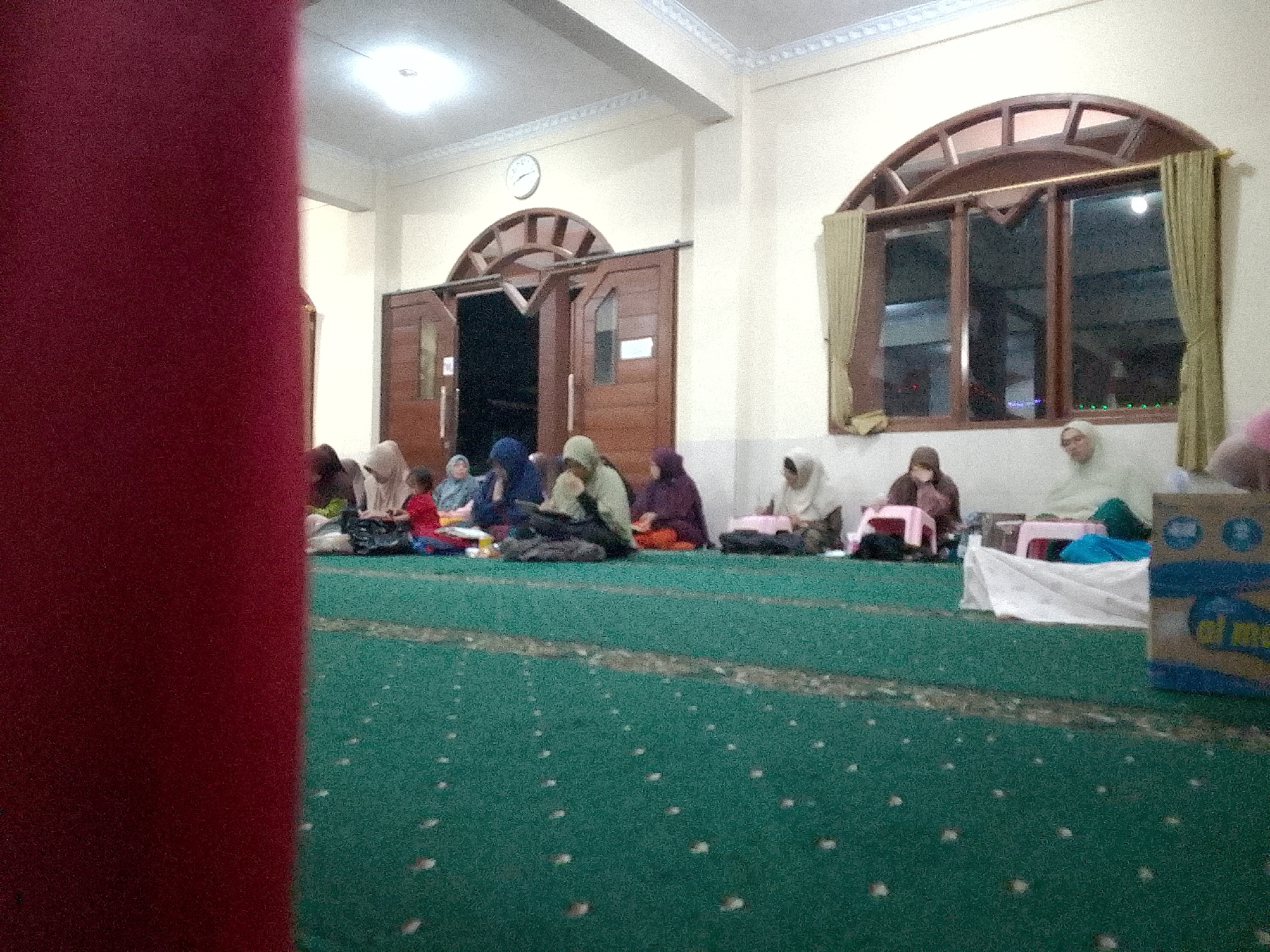 The image depicts a group of people, primarily women wearing hijabs, seated on a green carpeted floor inside a room. The room appears to be a place of worship or gathering, possibly a mosque, given the presence of a mihrab-like arched structure above a set of double doors and a large arched window with wooden framing and curtains. A clock is visible on the wall above the doors. Several individuals are engaged in prayer or quiet contemplation, with some appearing to read from books. Objects such as bags, a blue bucket, and a cardboard box with Arabic writing are also present in the scene. The lighting suggests it might be evening or an indoor space with artificial illumination. The overall atmosphere is peaceful and serene. The scene is likely located in Kertajaya, Indonesia, as per the provided context.