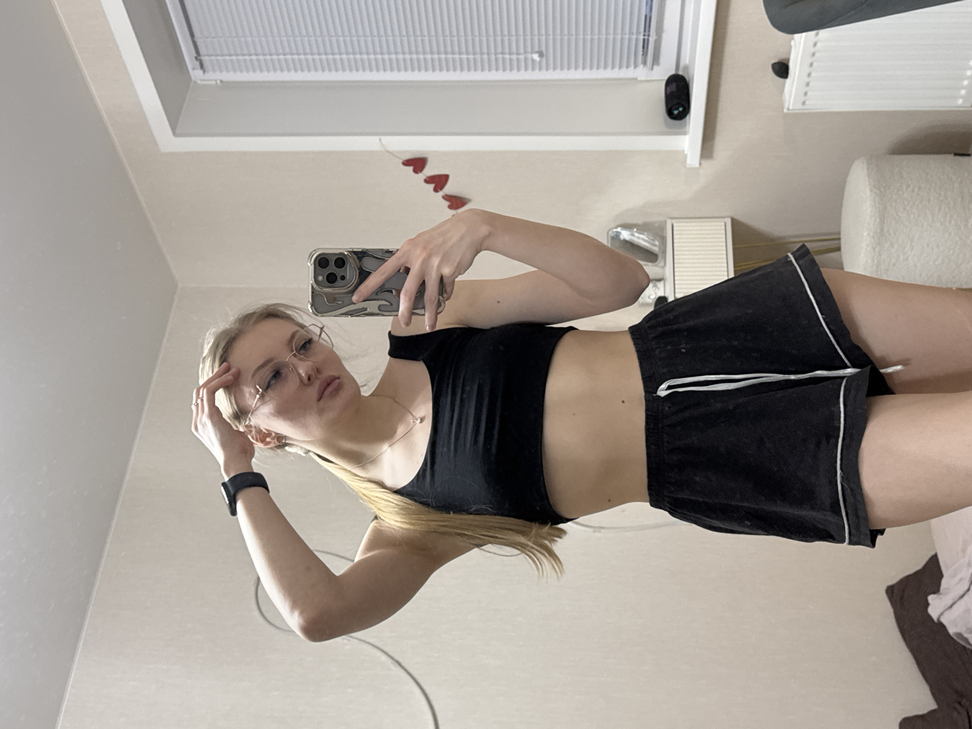 A woman in a black sports bra and black shorts with a white trim is taking a mirror selfie with a smartphone. The woman has blonde hair and wears glasses. There are red heart stickers on the wall near the top of the image. A window with blinds is in the background. Based on reverse image searches, the location is likely Perm, Russia.