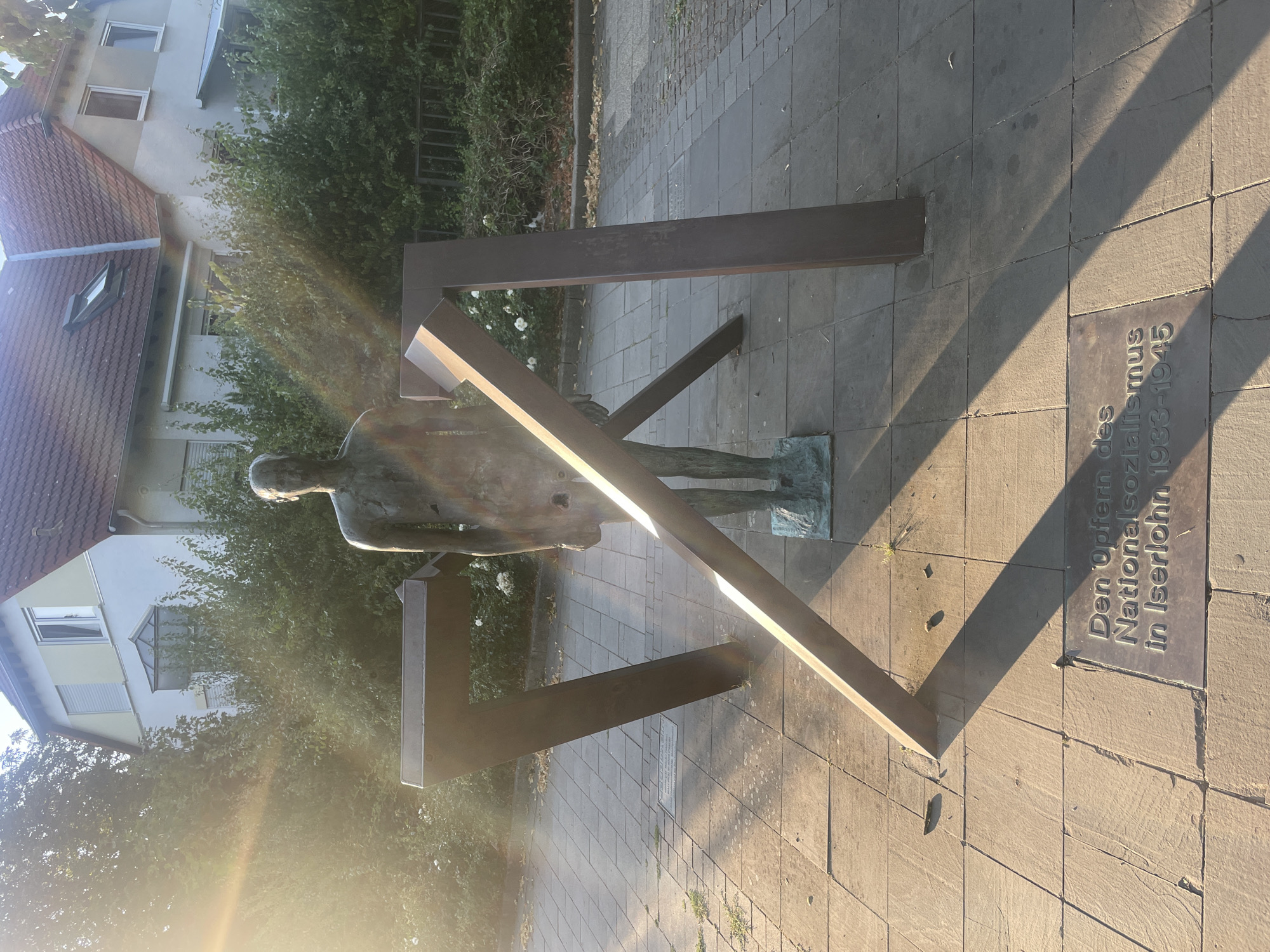 The image depicts a sculpture in Iserlohn, Germany, located outdoors on a paved walkway. The sculpture consists of a bronze statue of a full-bodied human figure with its head bowed, standing in front of and partially surrounded by three large, angular metal structures that form an abstract shape. A plaque on the ground in front of the sculpture is inscribed with German text. In the background, there is a two-story building with a red-tiled roof, surrounded by dense green foliage. Sunlight streams in from the left side of the frame, creating lens flare and casting long shadows across the scene. The ground is paved with square tiles, and some dry leaves are scattered on the path. The overall impression is of a memorial or public art installation.