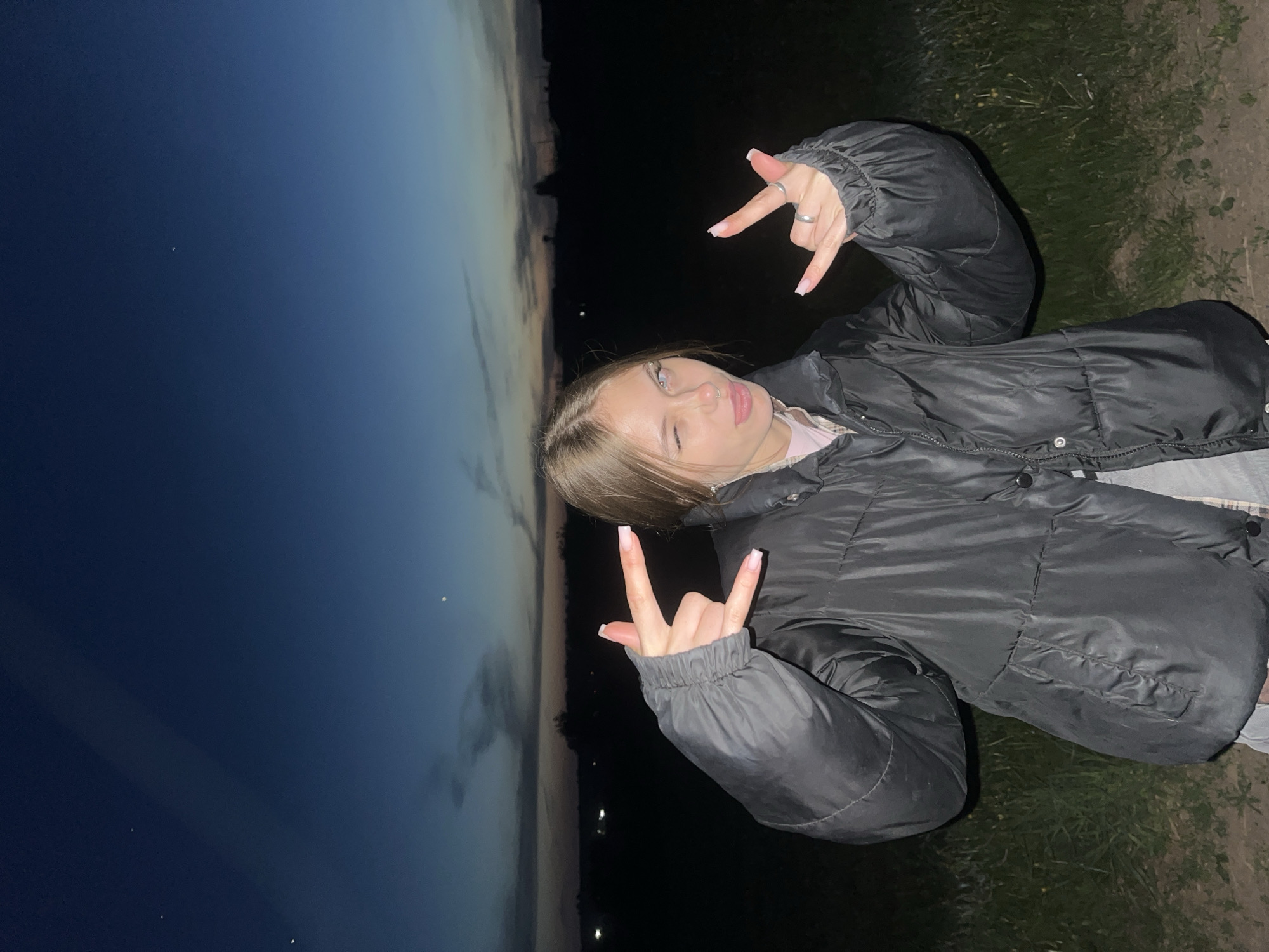 A young person is outdoors during twilight, likely near Фоки, Russia. The individual, facing the camera at an angle, has light brown hair, fair skin, and is wearing a black puffer jacket. They are making the "rock on" hand gesture with both hands, wearing multiple rings. To the left is a gradient sky transitioning from deep blue to a light orange-yellow horizon. The background behind the person appears to be a dark, grassy area with indistinct, possibly urban, lights visible near the horizon.