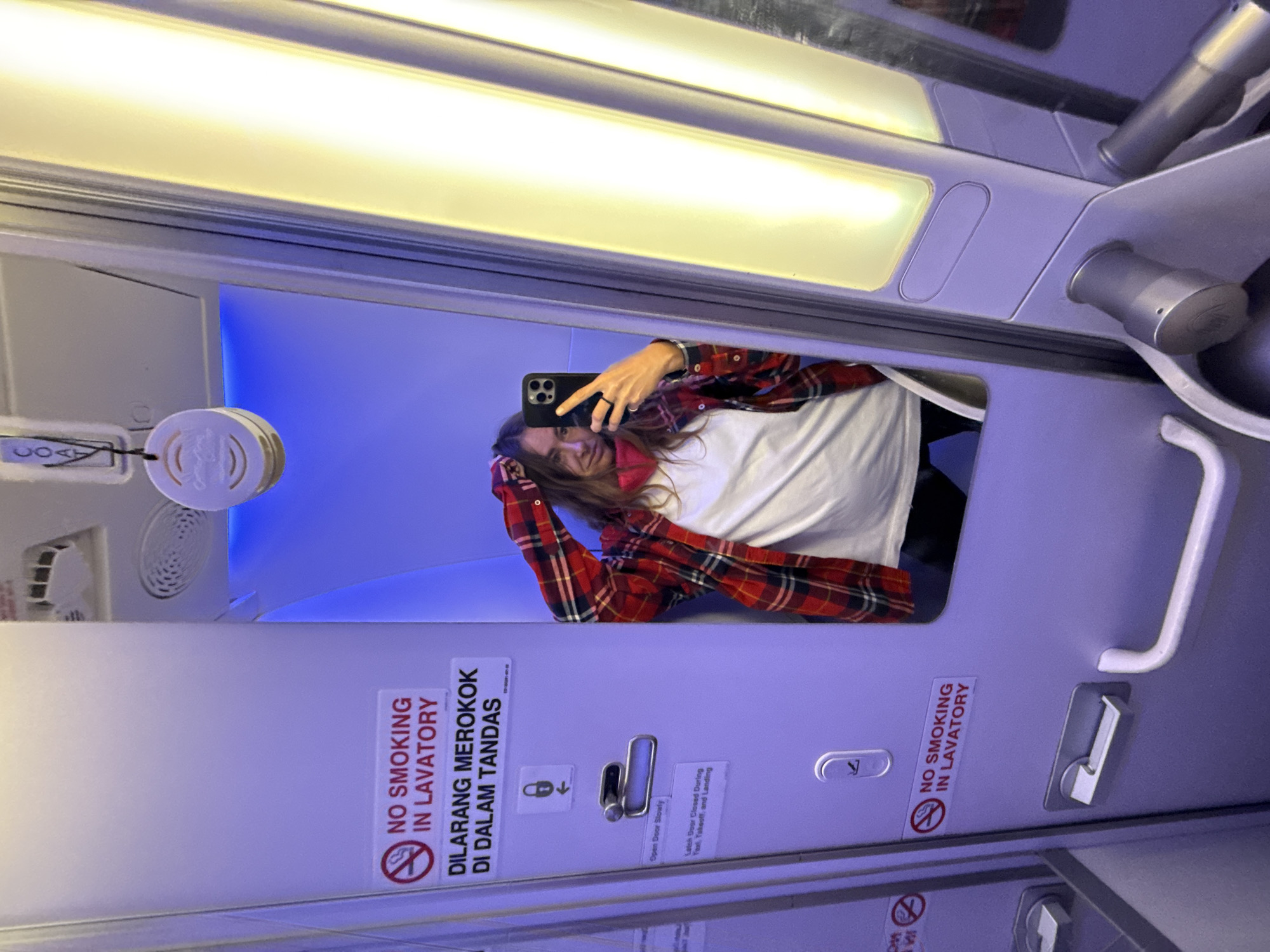 The image is an interior shot of an airplane lavatory, likely taken as a selfie reflected in the mirror. A person holding a smartphone is visible in the mirror's reflection. The lavatory door is white and has several signs: "NO SMOKING IN LAVATORY" is displayed with an image of a crossed-out cigarette and its Indonesian translation "DILARANG MEROKOK DI DALAM TANDAS." Symbols indicating "Open Door Slowly" and instructions to keep the latch door closed during taxi, takeoff, and landing are also present. A coat hook is visible above the door latch. The interior lighting is a mix of fluorescent white and blue.
The location is presumed to be Denpasar, Indonesia, inferred from the Indonesian language used in the "no smoking" sign.