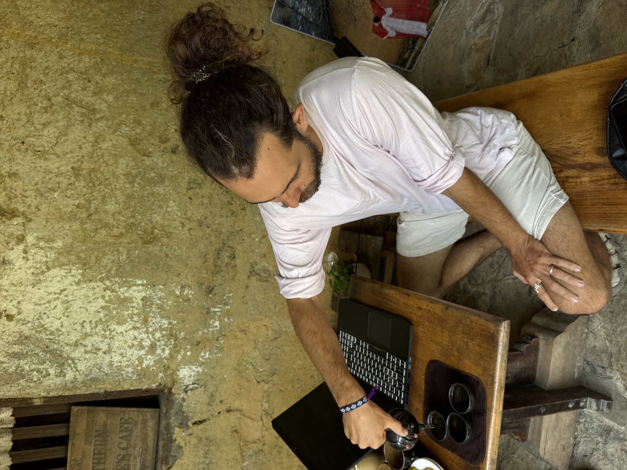 A man with a man bun is seated outdoors at a wooden table, working on a laptop. He is wearing a light pink shirt and white shorts. He is also wearing a bracelet on his left wrist and rings on both hands. He is pouring liquid from a small pot into a set of cups. The table also holds the laptop and a tray with the cups. The setting is outdoors with a rustic, worn-looking wall and floor. The location is Lũng Cú Commune, Vietnam.