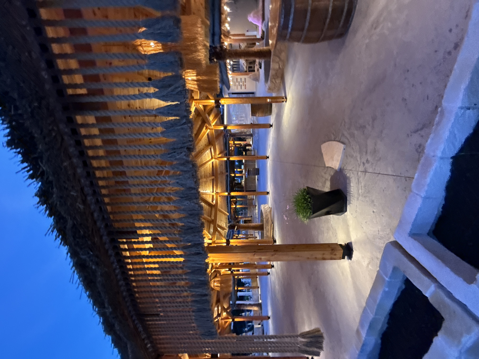 An outdoor setting at dusk, possibly a resort or entertainment venue, in Boreti, Montenegro, features a covered walkway or pavilion with a thatched roof supported by wooden posts. Rope decorations hang from the underside of the roof, with tassels visible at the ends. Warm lighting emanates from the interior, illuminating the wooden structure and the concrete ground. A potted green plant sits in a dark, angular planter on the ground. In the background, other covered areas with similar architectural elements are visible, along with what appear to be bar areas or seating arrangements. A large wooden barrel is situated on the right side of the frame. The sky above is a deep blue, indicating the transition from day to night.