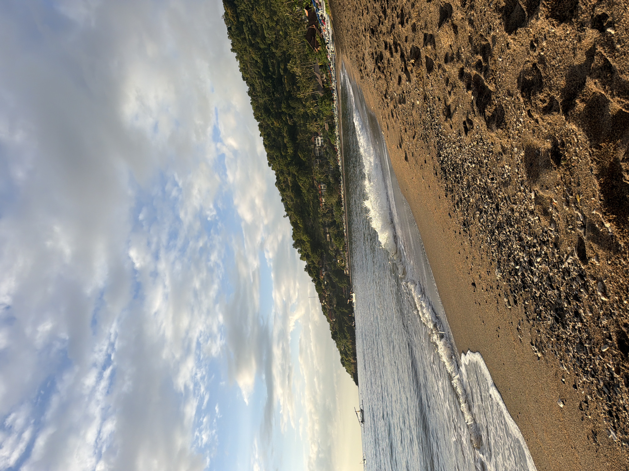 The image shows a beach scene in Abang, Indonesia. The foreground is dominated by golden sand with numerous footprints, interspersed with small pebbles. A gentle wave is breaking on the shore, creating white foam. In the middle ground, the calm ocean stretches towards a verdant hillside covered in dense trees and vegetation. Along the base of the hill, several buildings, likely resorts or homes, are visible, with some red-tiled roofs. Scattered along the coastline are several traditional outrigger boats, some with sails. The sky above is a mix of blue and white clouds, with the warm glow of late afternoon or early morning sun casting long shadows on the sand.