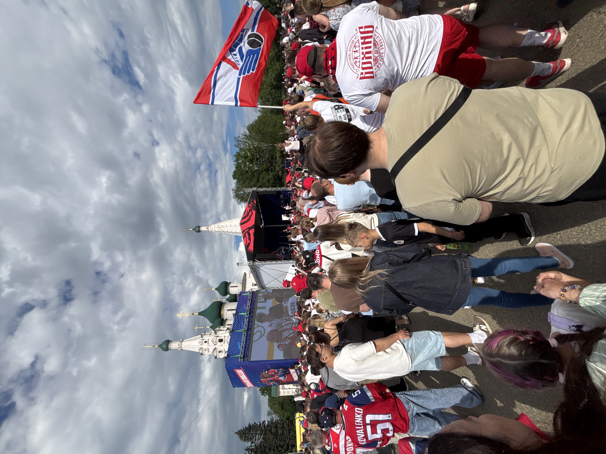 The image captures a scene in Rusanovo, Russia, where a large crowd has gathered to watch a hockey game on a large screen. In the foreground, people are standing and sitting, some wearing hockey jerseys and clothing with the "Lokomotiv" logo. A red and white flag with a blue hand logo is visible. A man wears a white shirt with the words "BOXING" and "TRADITIONAL RUSSIAN". In the background, a white cathedral with green domes stands, contrasting with the cloudy sky. The screen displays a hockey scene with the year "2026" printed on it.