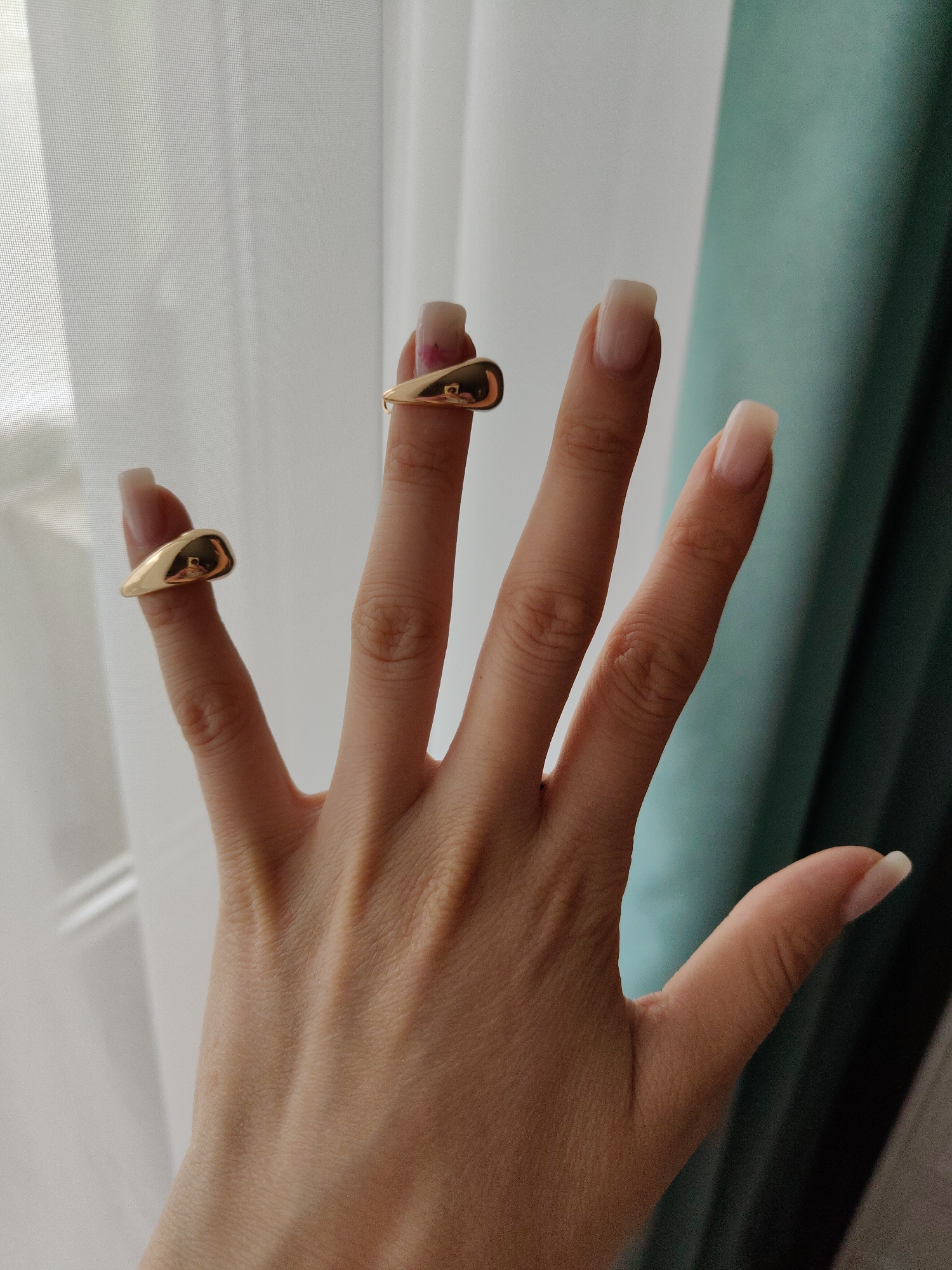 A pale hand displays two gold rings on the index and thumb of the hand. The hand is set against a backdrop of a white curtain, as well as a green curtain. There is a small pink mark on the finger. The location and city are unknown.