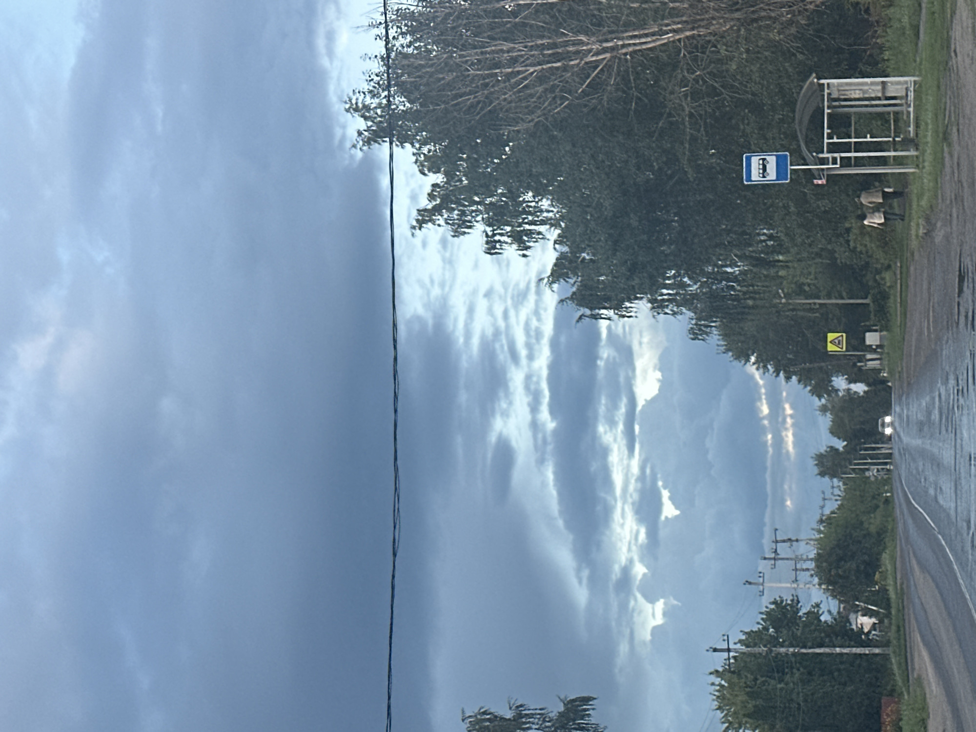 The image shows a cloudy sky over a road in Rasskazovo, Russia. There are trees on both sides of the road, and a bus stop with two people walking away from it. The road is wet, suggesting it has recently rained. In the distance, a car is approaching, and there are power lines running along the road.