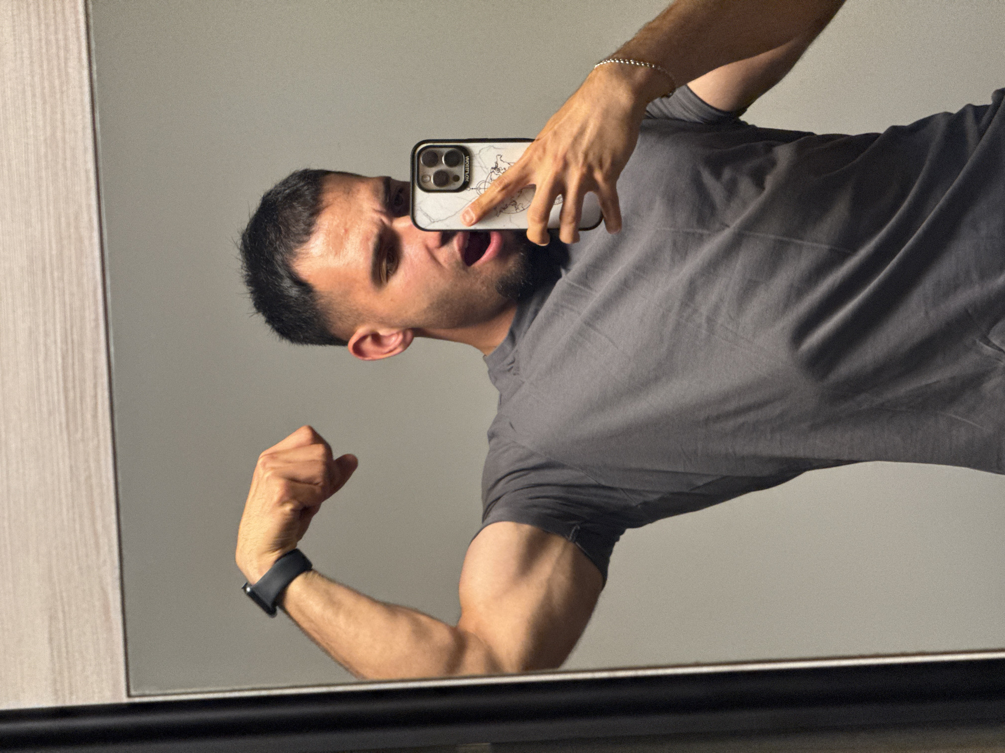 A man is posing for a selfie in a bathroom. He is wearing a gray t-shirt and flexing his bicep. He is holding his phone in his right hand, which is also wearing a silver bracelet. The man has short dark hair and a light beard. He is looking into a mirror, and his mouth is open. The background is a light gray wall. There are no specific details that indicate the location in the city of Shumen, Bulgaria.