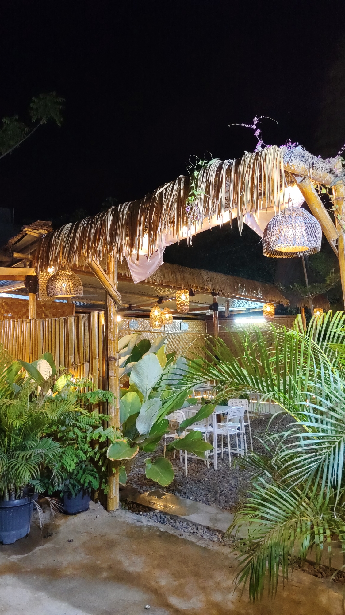 The image depicts a tropical outdoor dining area at night, likely a restaurant or cafe, in Keudebayu, Indonesia. The scene is illuminated by hanging woven lamps and string lights, casting a warm glow on the surroundings. The structure features a thatched roof supported by bamboo posts, with walls made of woven bamboo. Lush green tropical plants, including large-leafed plants and palm fronds, are arranged around the dining area, creating a natural and inviting atmosphere. White tables and chairs are set on a gravel ground, suggesting a seating arrangement for patrons. The overall ambiance is relaxed and exotic, characteristic of Southeast Asian outdoor venues.