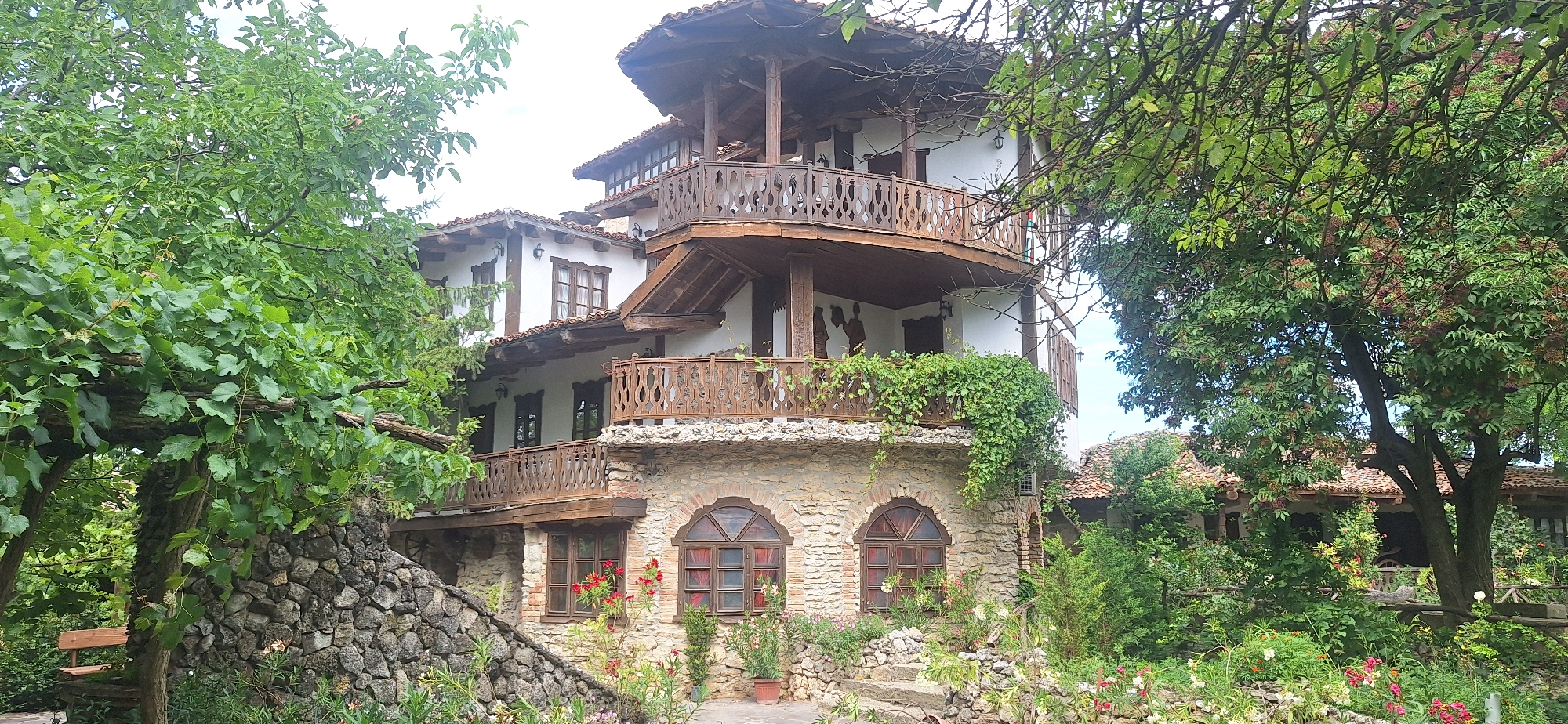 The image shows a multi-story building with traditional architecture, likely a guesthouse or restaurant, nestled in a lush, green setting. The building features white stucco walls, terracotta tiled roofs, and intricate wooden balconies with decorative railings. The lower levels are constructed with stone. There are numerous windows, some with arched tops. Greenery, including trees, vines, and flowers, surrounds the building, with a stone wall and a wooden bench visible in the foreground. The scene evokes a rustic and inviting atmosphere, suggesting a peaceful rural location. This building is located in Kalimantsi, Bulgaria.