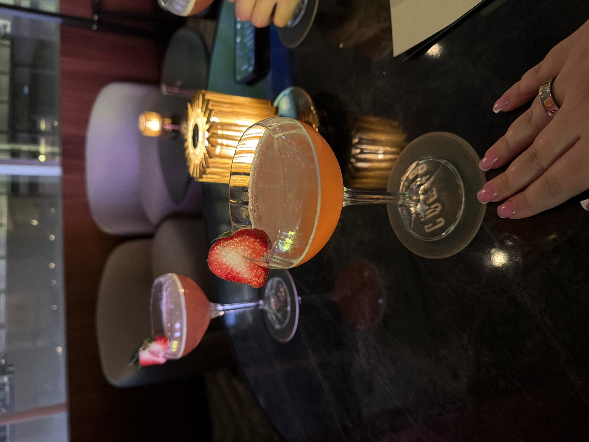 Two vibrant cocktails are the central focus of this indoor scene, set on a dark, reflective tabletop. The most prominent cocktail, positioned in the center-right, is an orange-hued drink served in a coupe glass, elegantly garnished with a fresh, halved strawberry hooked onto its rim. To the left and slightly behind it, another cocktail, pink in color and also in a coupe glass, is visible with a smaller strawberry slice as garnish.
A person's hand, adorned with light pink French-tip manicured nails and a silver band ring on the ring finger, rests casually on the dark table surface to the right of the orange drink. Further in the blurred background, another hand can be seen holding a glass, indicating at least two individuals are present.
Behind the orange cocktail, a decorative, golden-colored, metallic object, possibly a small lamp or candle holder with a ribbed surface, emits a warm glow, contributing to the ambient lighting. The dark, glossy table reflects parts of the glasses and lights. The background is softly blurred but suggests an upscale indoor setting with muted colors of furniture, possibly plush seating or booths, and additional warm lights.
The activity taking place appears to be a relaxed social occasion, likely an evening or nighttime gathering at a bar, lounge, or restaurant in New York, United States of America. The overall atmosphere is sophisticated and inviting. On the base of the orange cocktail glass, the partial text "CUE" is visible.