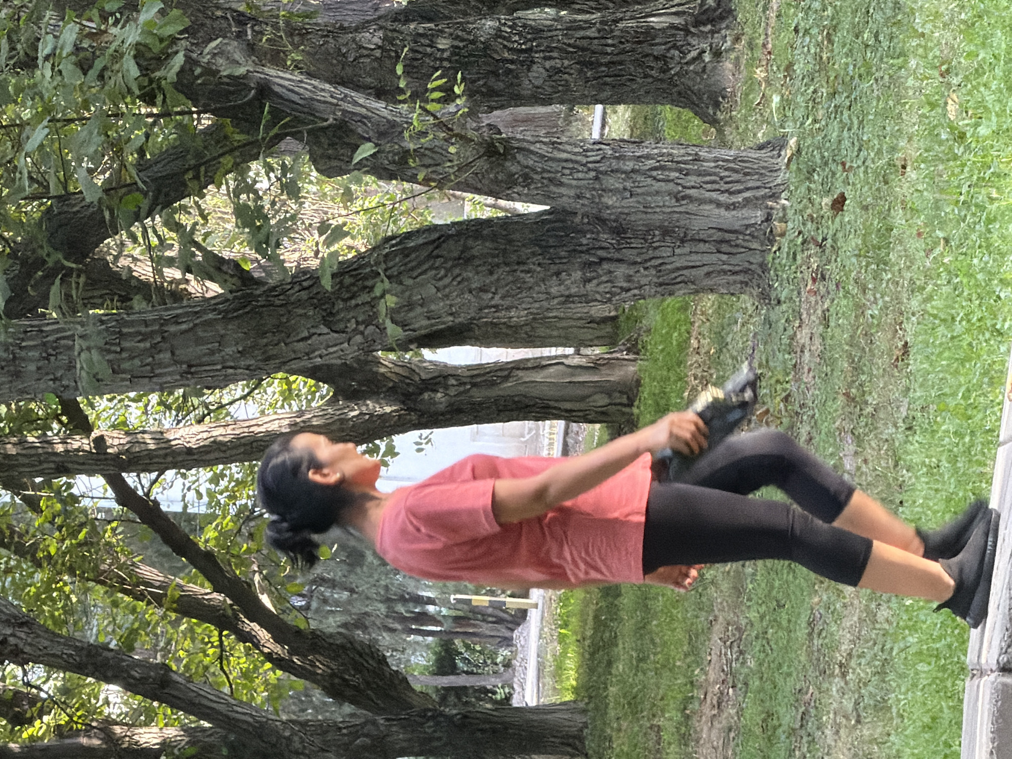 A woman with dark hair pulled back in a bun walks from left to right in a park setting. She is wearing a short-sleeved coral t-shirt and black capri-length leggings. On her feet, she has on black athletic shoes. She is holding a dark-colored cylindrical object in her right hand. The ground is covered in green grass and some patchy brown leaves. She is walking along a light gray concrete curb. Behind her, there are several large trees with textured bark, and foliage overhead. In the background, there is a lighter building structure and more trees. The lighting suggests it is daytime, with sunlight filtering through the leaves.