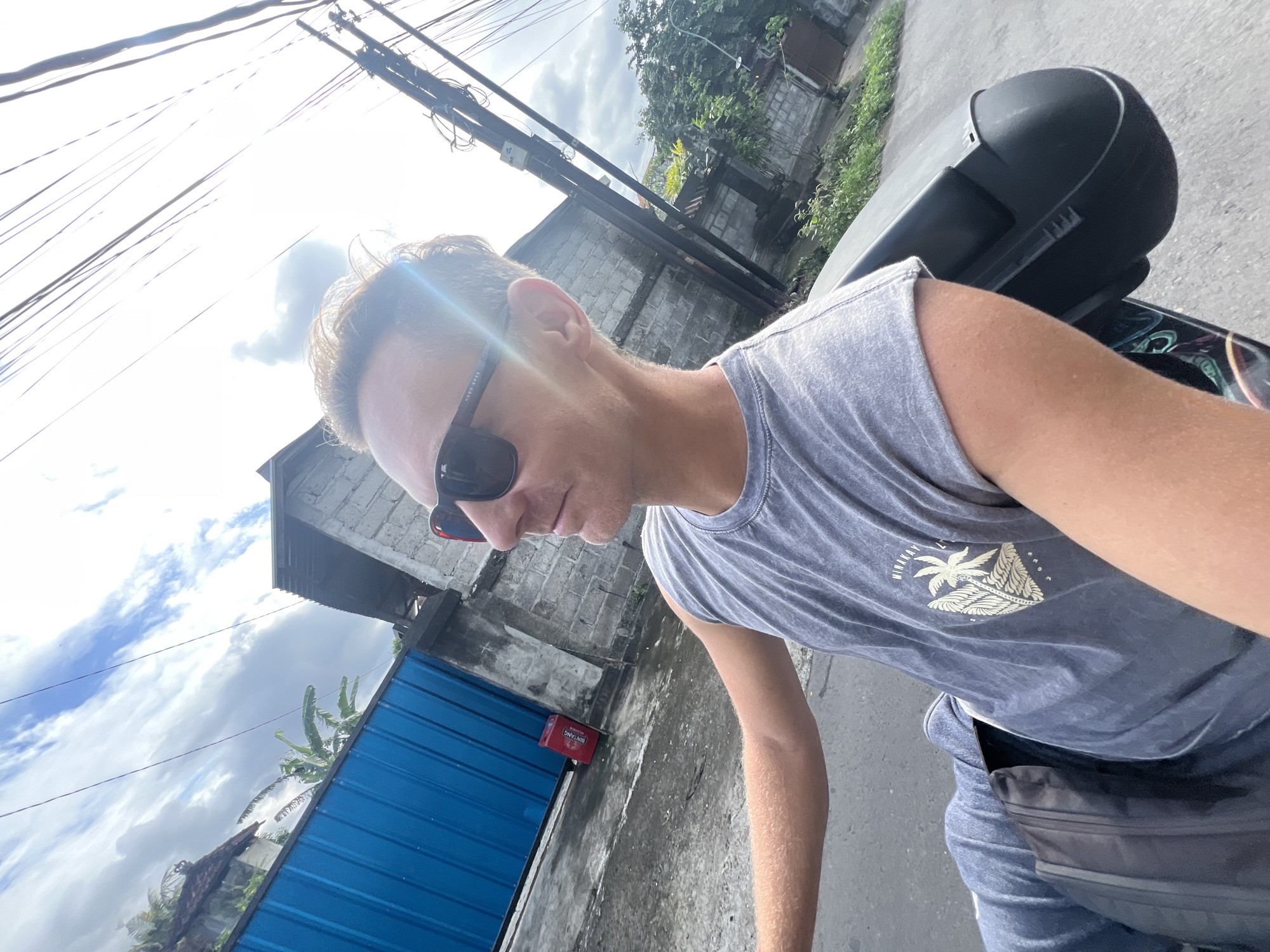 A person wearing sunglasses and a gray t-shirt with a palm tree design is on a motorcycle. The person is in Sukawati, Indonesia. A blue metal fence, a building with a gray brick facade, and overhead power lines are visible. A black storage box is on the motorcycle behind the person.